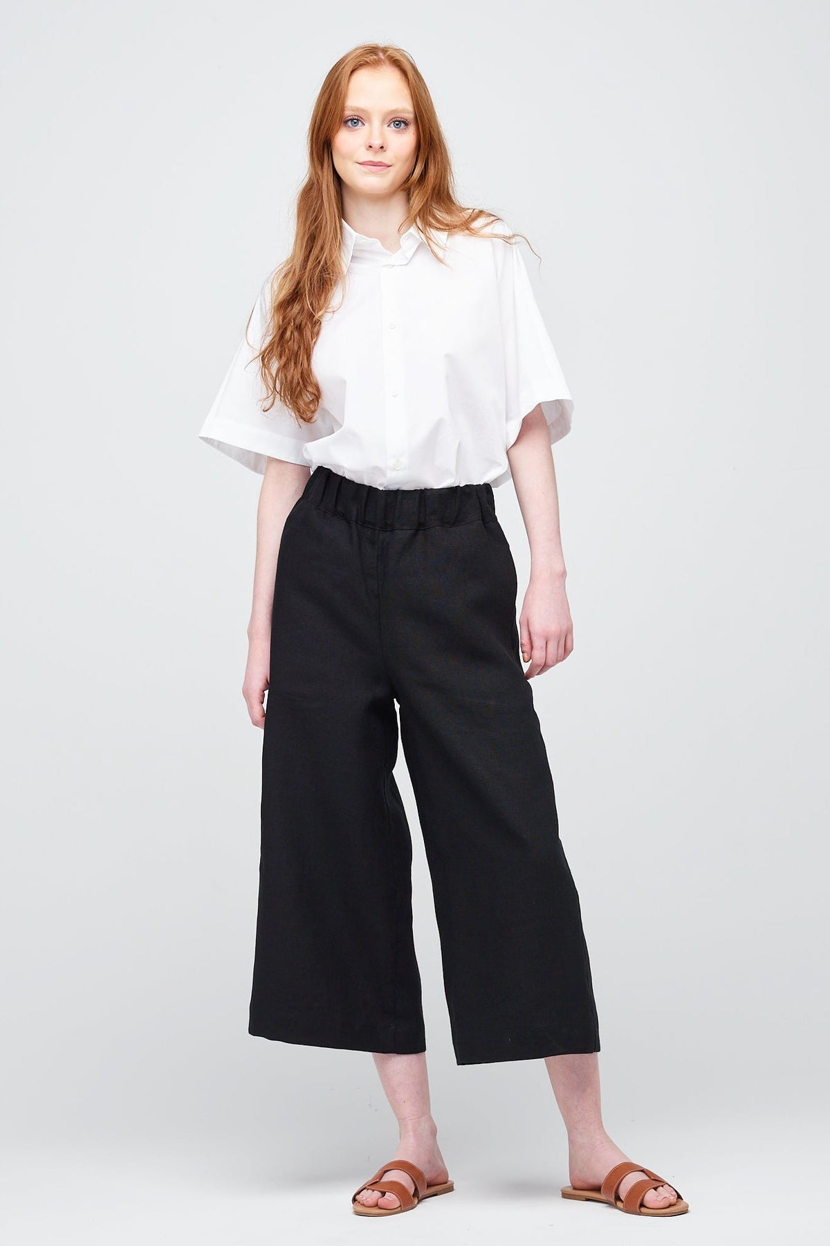 A young white women with ginger hair wearing short pj trouser in black linen, styled with an oversized white shirt and brown sandal.