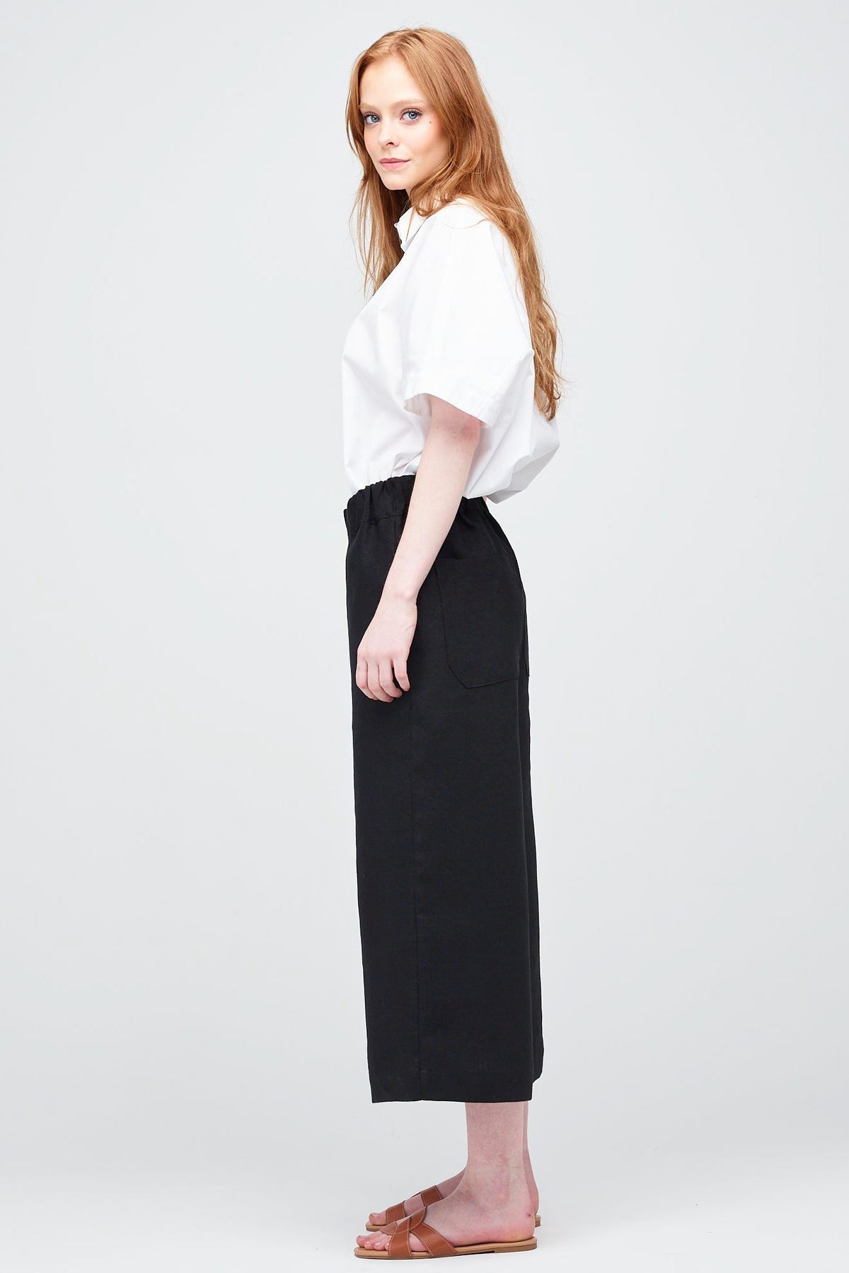 Side shot of a young white women with ginger hair wearing short pj trouser in black linen, styled with an oversized white shirt and brown sandal.