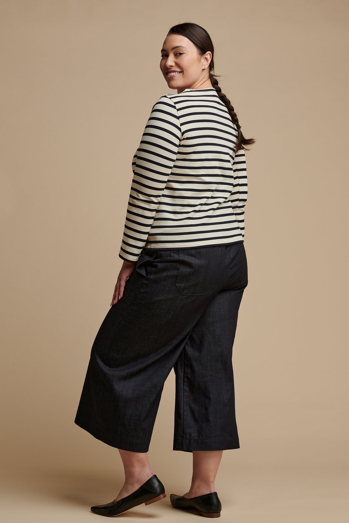 Smiling female with brown hair tied back, wearing indigo denim short pj pants, facing away from the camera and looking back over her shoulder.