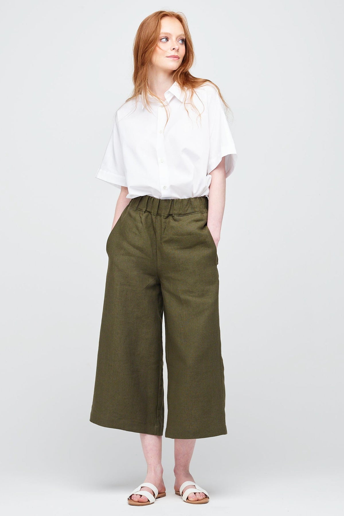 A young white women with ginger hair wearing short pj trouser in a khaki coloured linen, styled with an oversized white shirt and sandal.