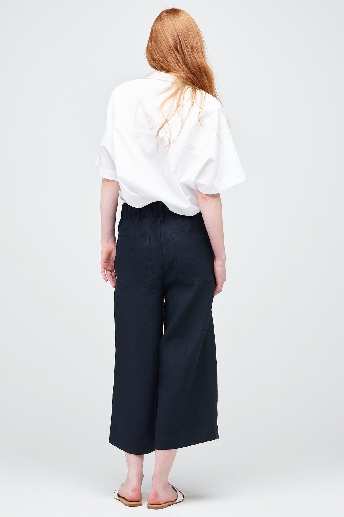 Rear shot of a young white women with ginger hair wearing short pj trouser in navy linen, styled with an oversized white shirt and sandal.
