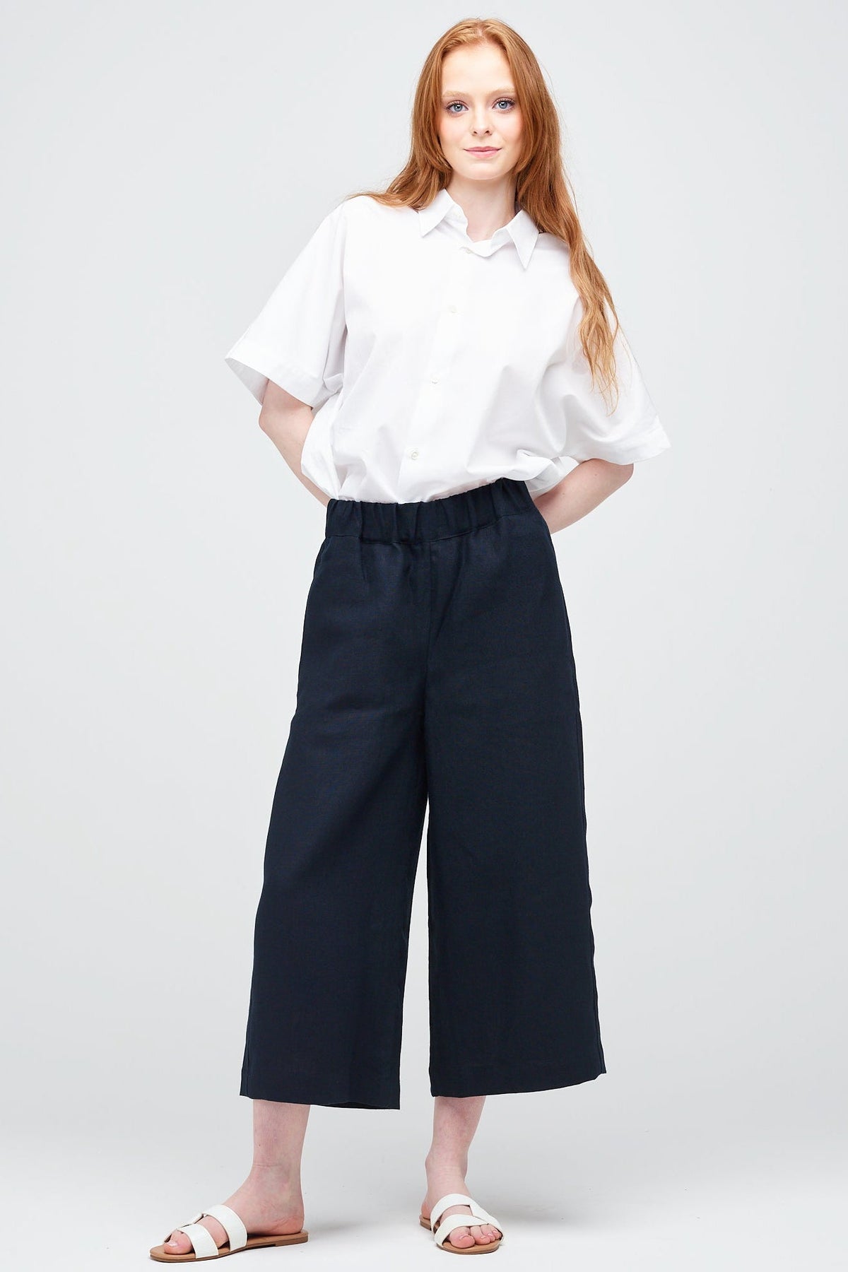 A young white women with ginger hair wearing short pj trouser in navy linen, styled with an oversized white shirt and sandal.
