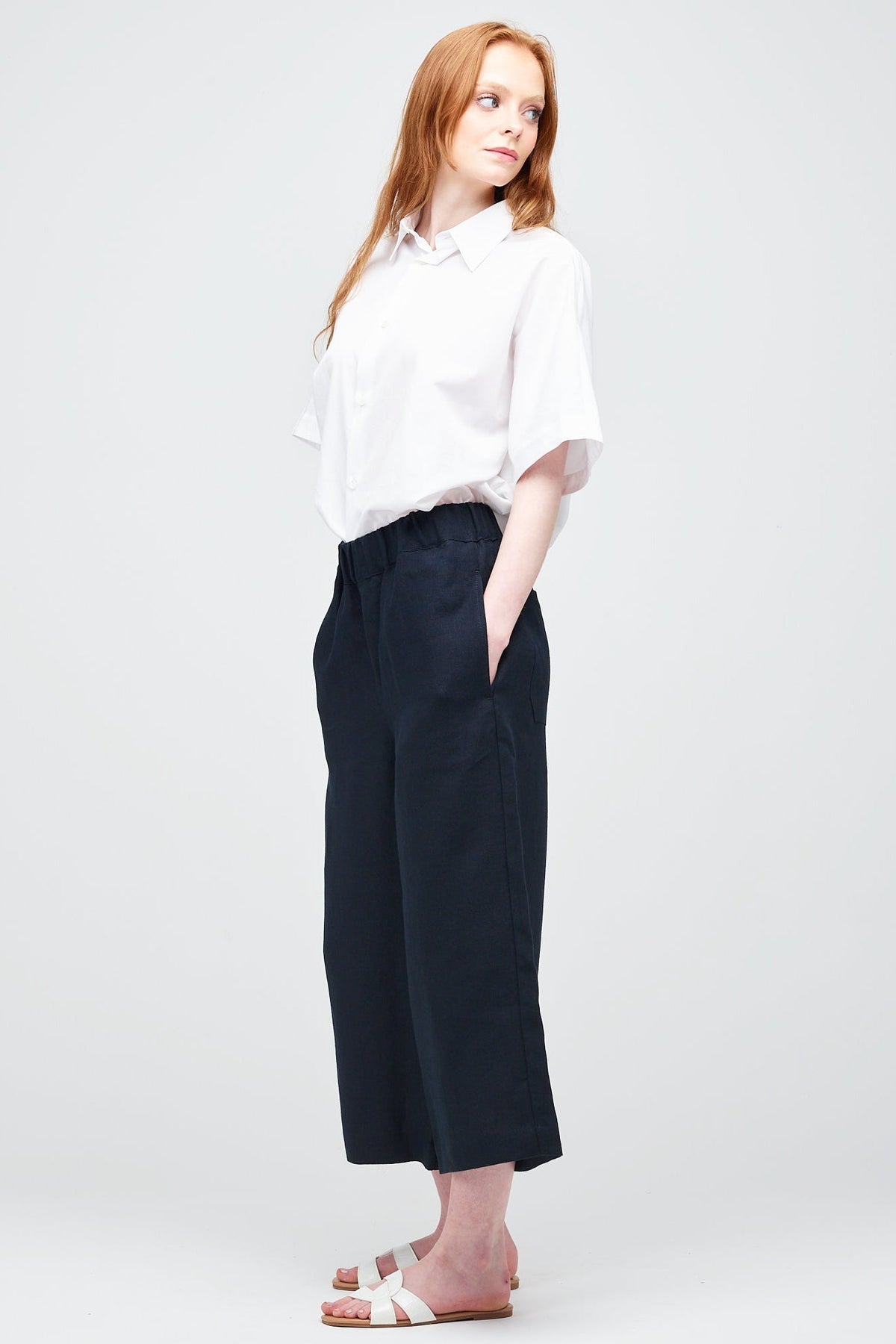 Side shot of a young white women with ginger hair wearing short pj trouser in navy linen, styled with an oversized white shirt and sandal.