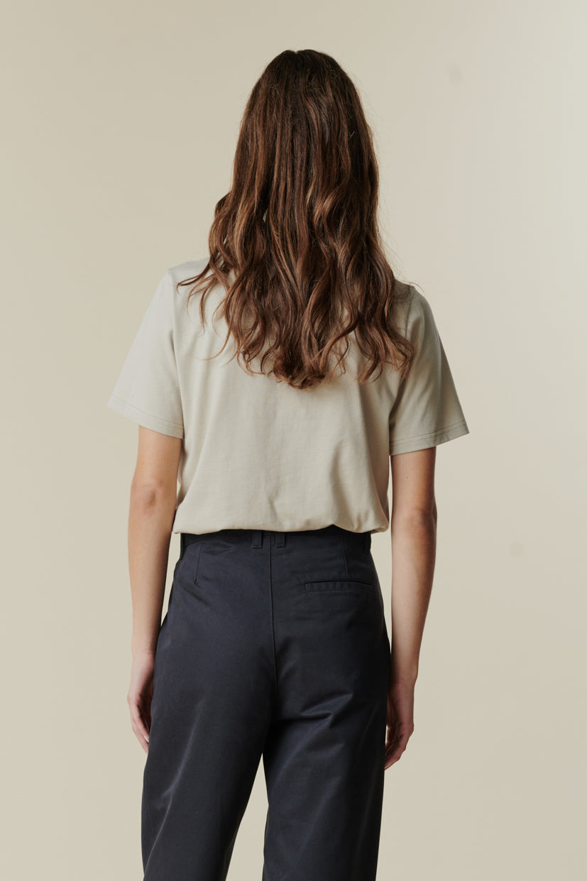 Thigh up image of the back of brunette female wearing short sleeve t shirt in stone paired with pleated trousers in navy