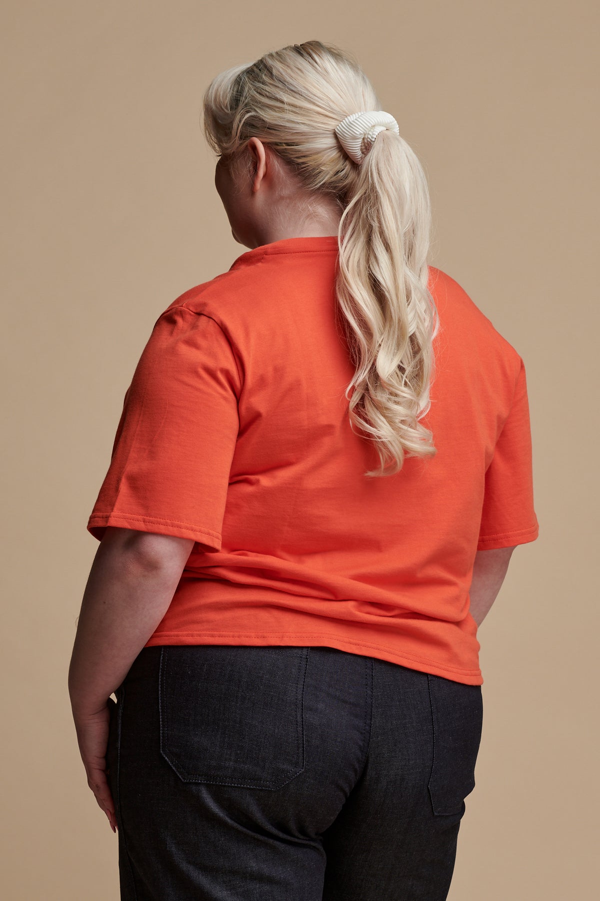 Thigh up of the back of female with blonde hair tied back into a ponytail wearing short sleeve v neck t shirt in flame, paired with work trousers in denim.