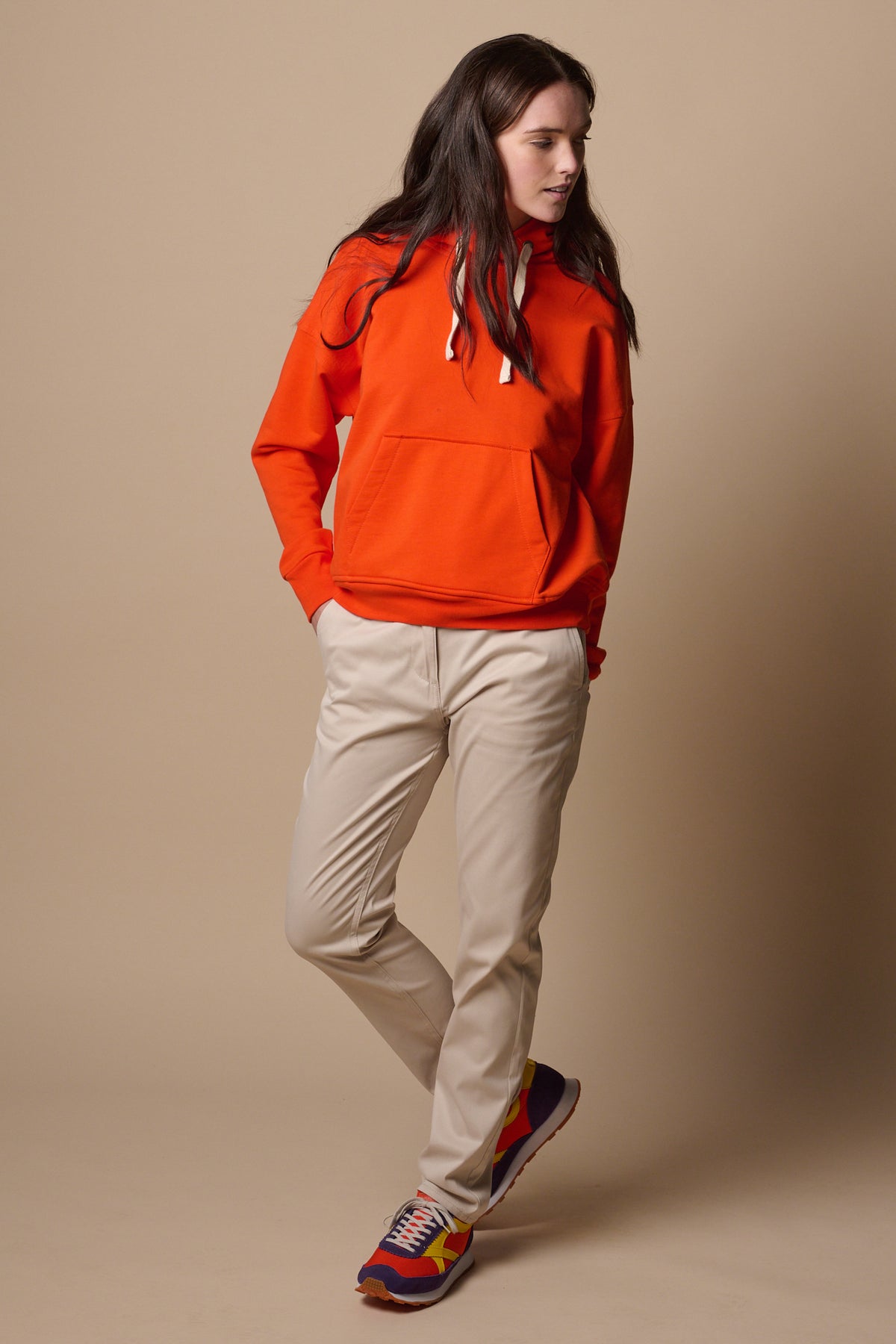 Full body image of brunette female wearing slim stretch chinos in light stone paired with hooded sweatshirt in flame red