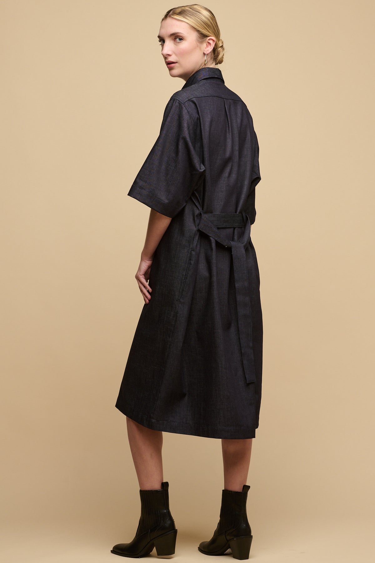 Thigh up image of female wearing straight denim dress in indigo, with the wait belt tie tied at the back. Worn with heeled black boots