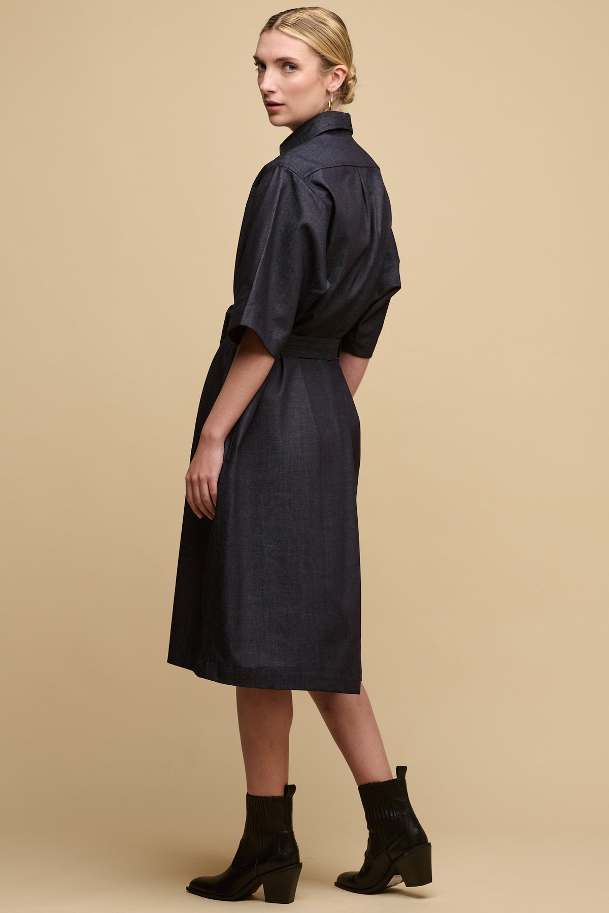 Back of female wearing straight denim dress in indigo with waist belt tied to synch the waist, paired with black heels boots