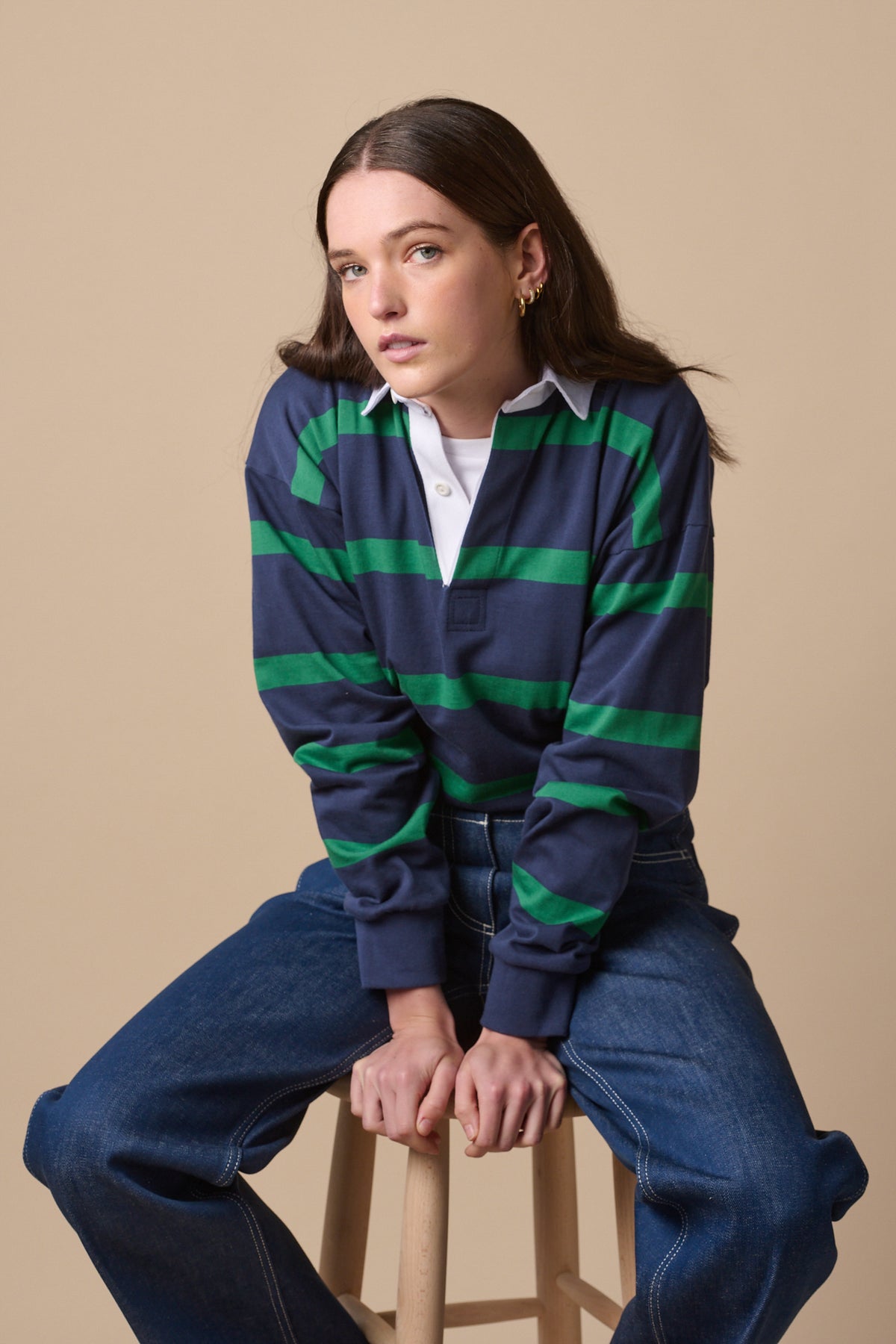 Female wearing striped rugby shirt in navy emerald, posing sat on stool.