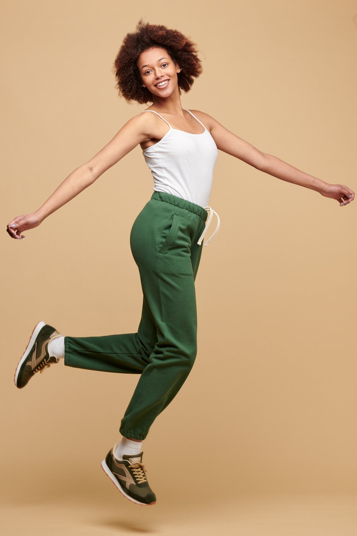 Female jumping in the air with her arms wide wearing Community Clothing 100% cotton sweatpants in bottle green paired with white camisole tucked into sweatpants and Walsh Footwear X Community Clothing Beacon trainer in olive/beige