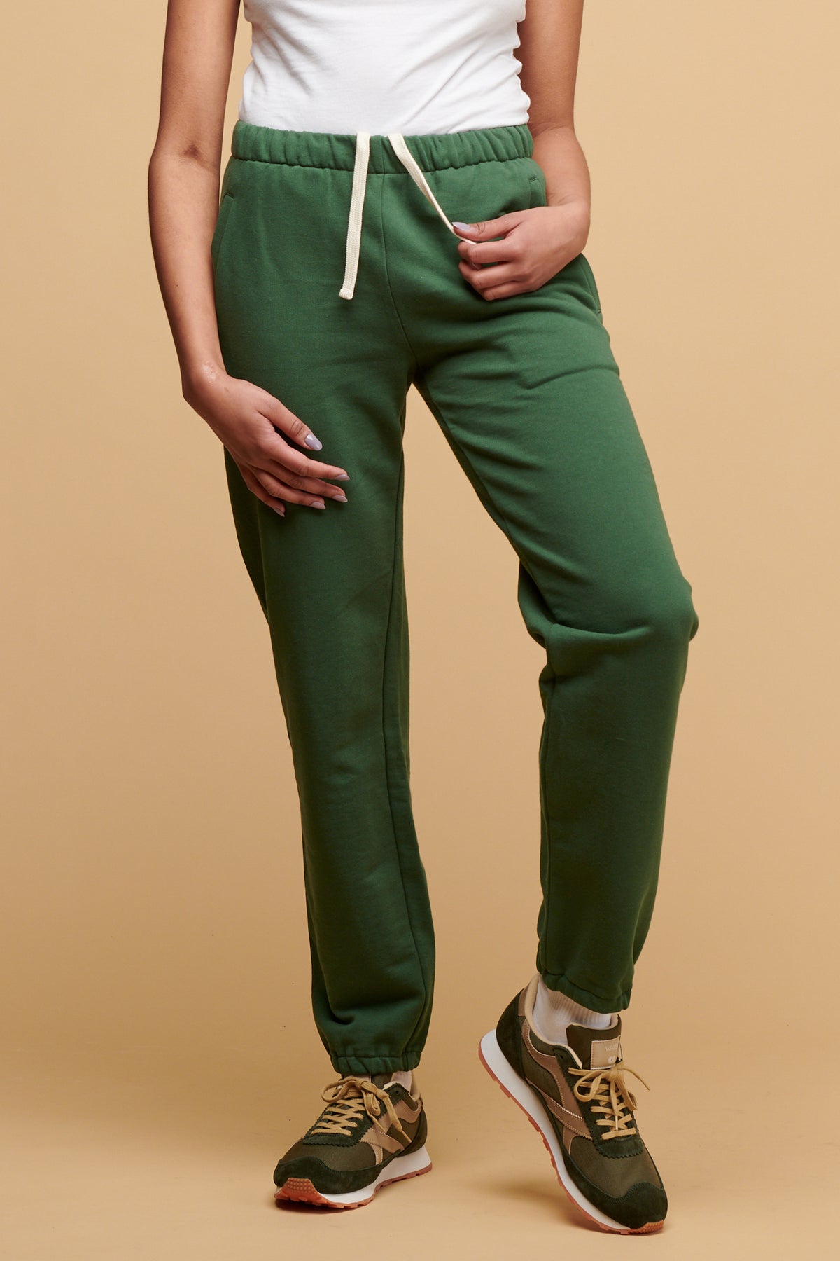 Chest down of female wearing Community Clothing 100% cotton sweatpants in bottle green with ecru drawcord hanging out paired with white camisole tucked into sweatpants and Walsh Footwear X Community Clothing Beacon trainer in olive/beige