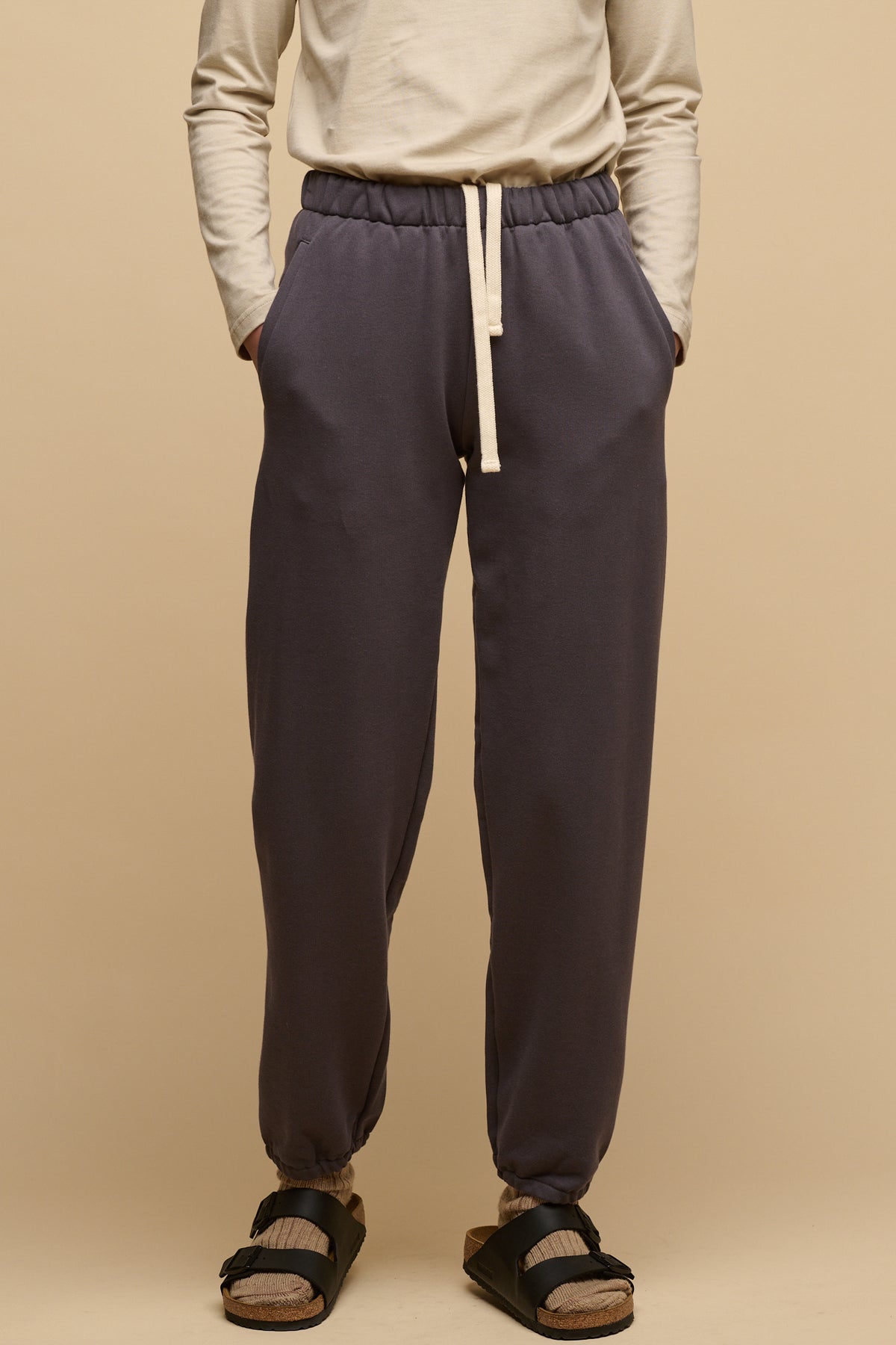 Waist down image of female wearing sweatpants in slate grey with hands in front pockets and ecru drawstring for waist out at the front. Worn with long sleeve t shirt in stone tucked in and cushioned wool walking socks in fawn