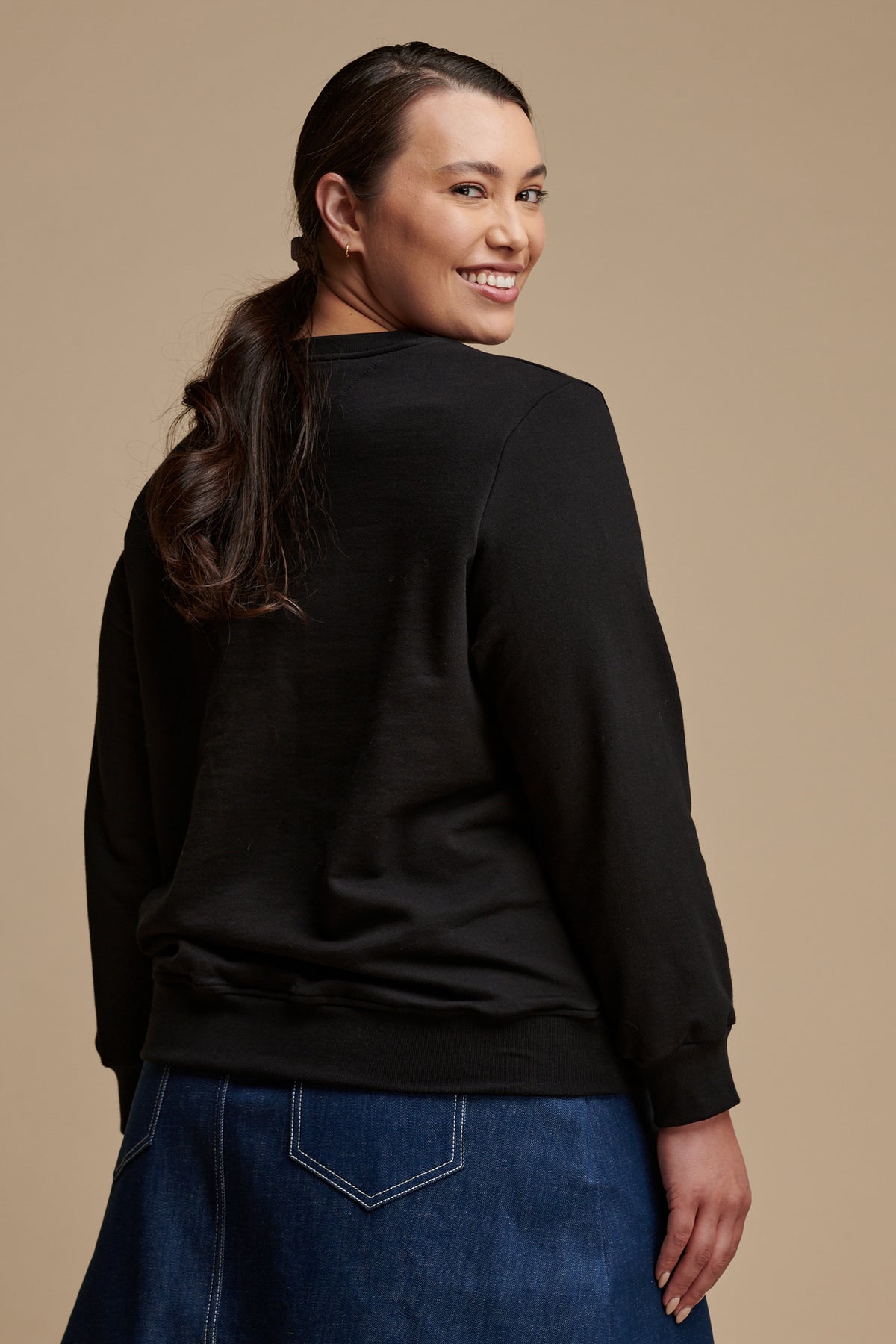 Thigh up of the back of female looking back over her shoulder smiling to the camera wearing sweatshirt in black paired with bobbie skirt blue.