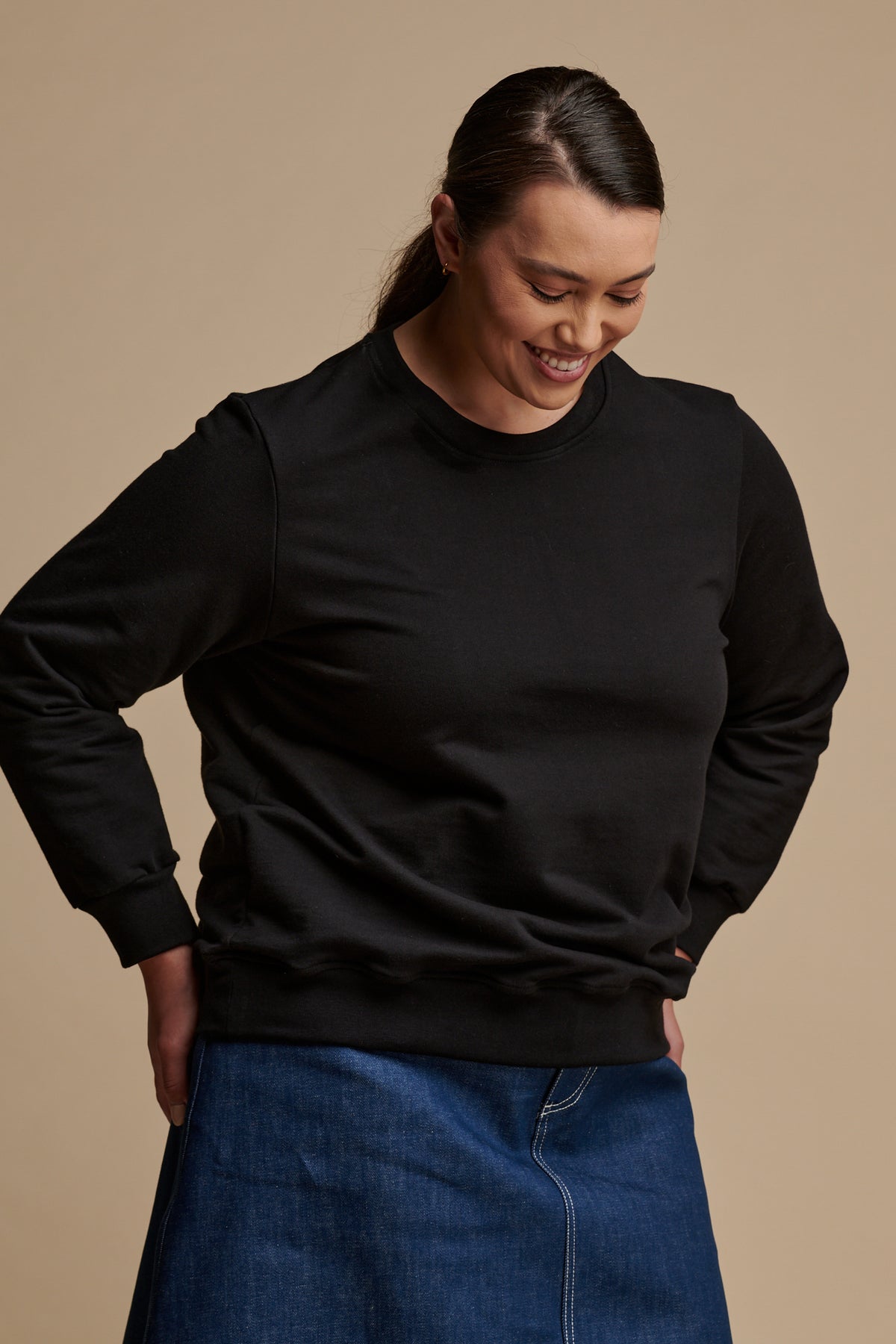 Thigh up of smiley female looking at the ground wearing sweatshirt in black with her hands on the back of her hips.