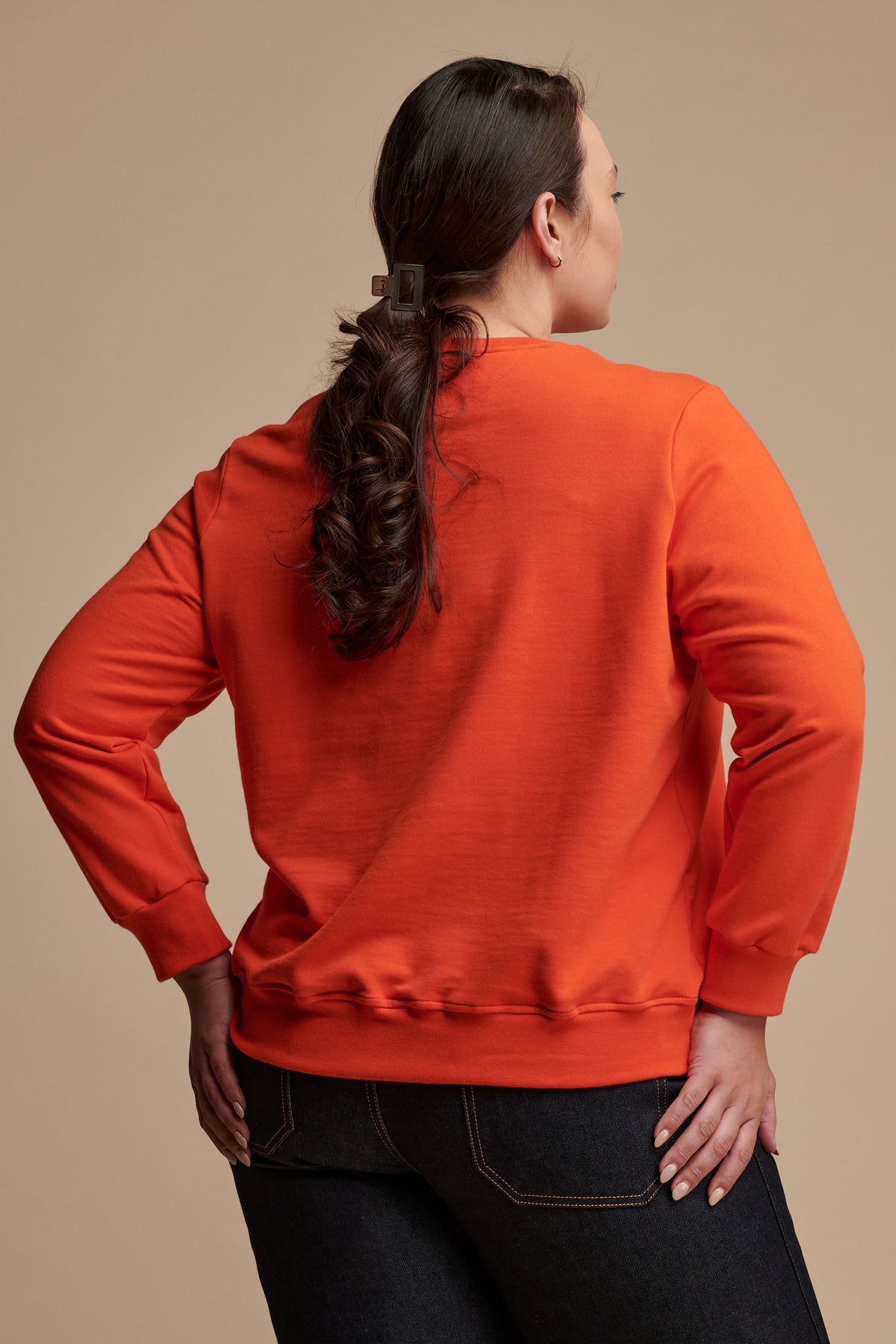 Thigh up of the back of female with dark hair tied back wearing sweatshirt in flame red paired with work jeans in indigo.