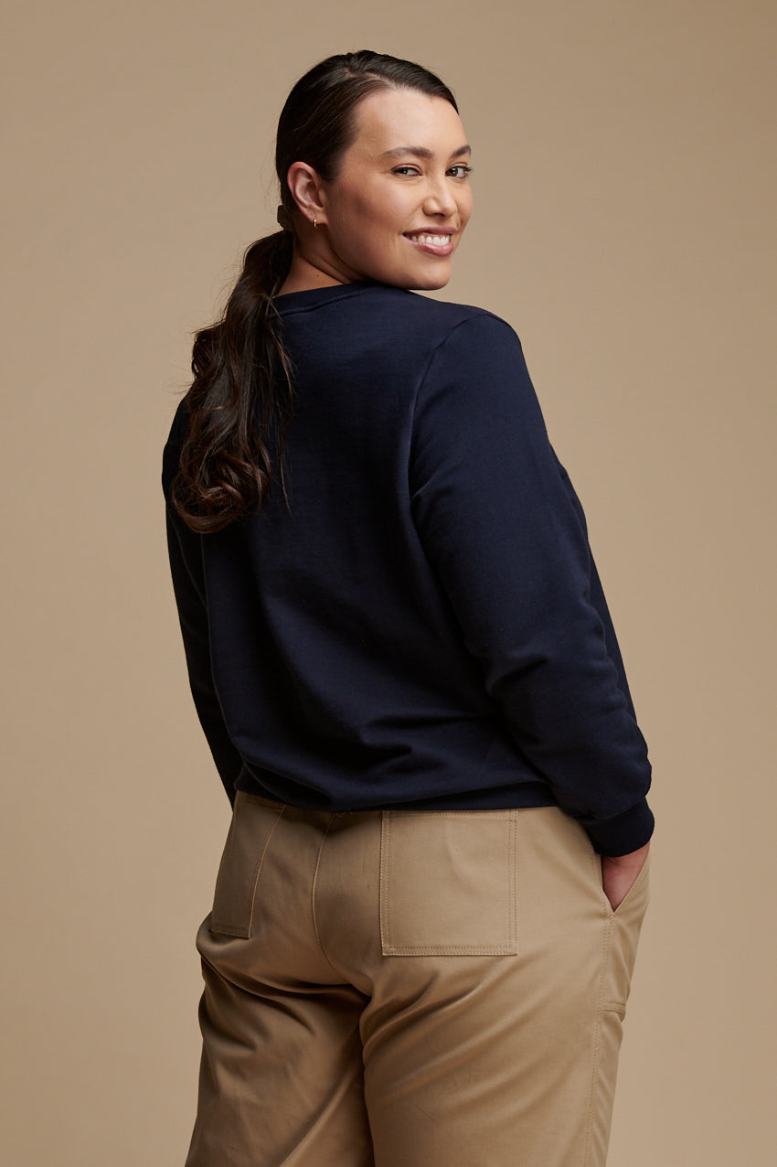 Thigh up of the back of female looking back over her shoulder smiling to the camera wearing sweatshirt in navy paired with camerawoman pants in putty.