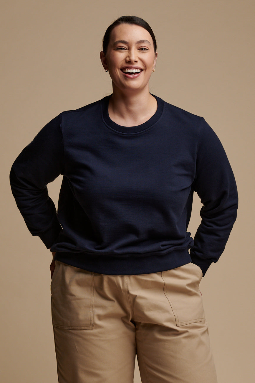 Thigh up of smiley female wearing sweatshirt in navy paired with camerawoman pants in putty.