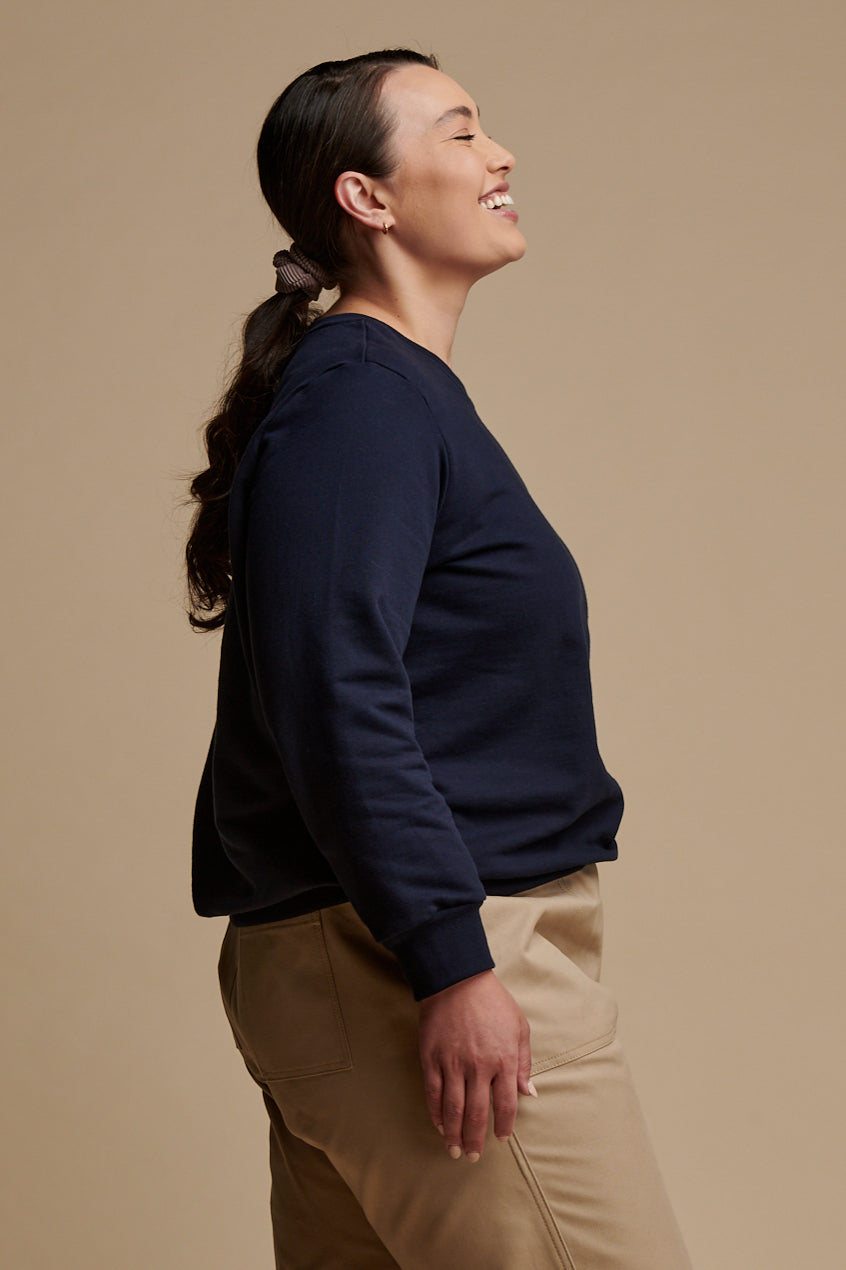 Thigh up of the side of smiley female wearing sweatshirt in navy paired with camerawoman pants in putty.
