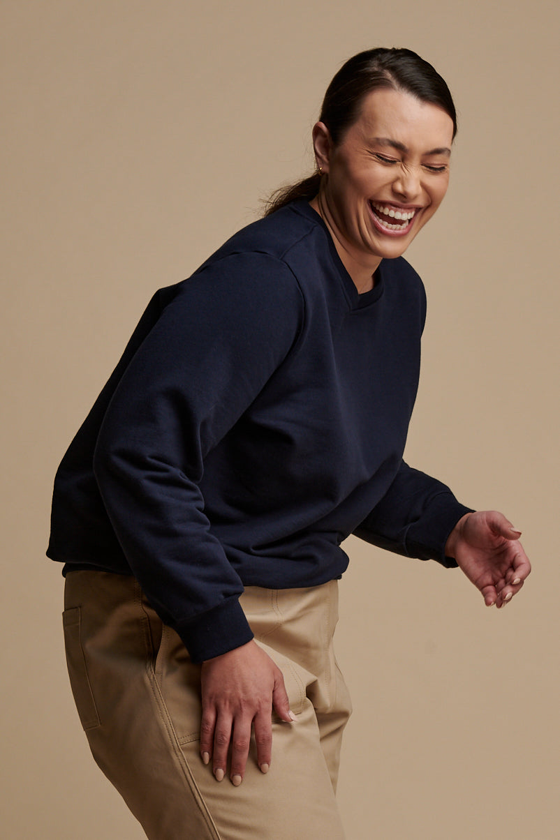 Thigh up of female leaning forward and laughing wearing sweatshirt in navy paired with camerawoman pants in putty.