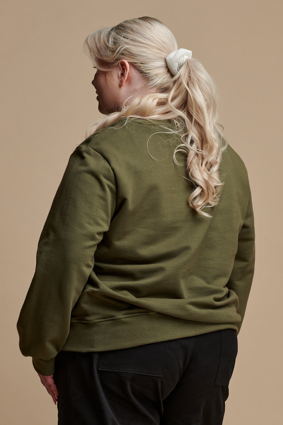Thigh up of the back of female with long blonde hair tied back into a ponytail wearing sweatshirt in olive paired with camerawoman pant in black.