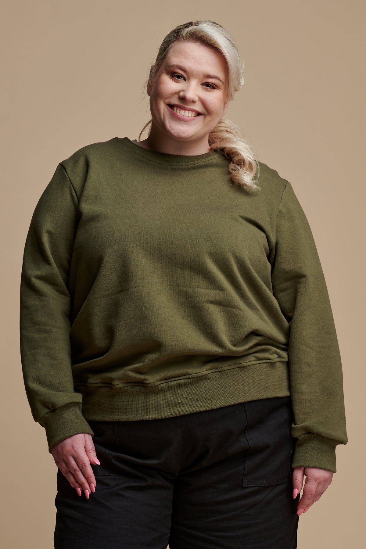 Thigh up of female with blonde hair tied back wearing sweatshirt in olive paired with camerawoman pants in black.