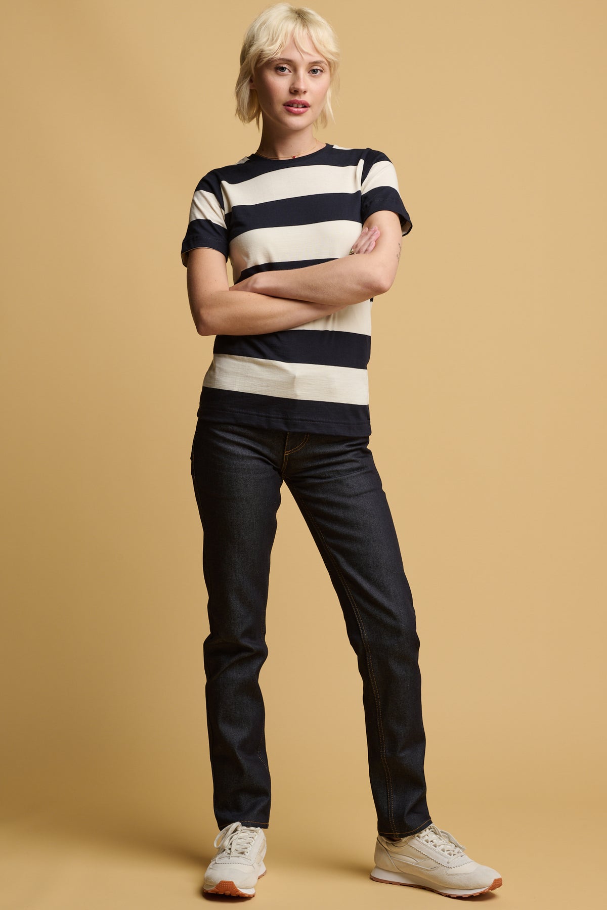 Thigh up image of female with her arms crossed wearing short sleeve wide stripe t shirt in ecru navy paired with straight leg jeans in indigo and ivory and beige parlick trainers