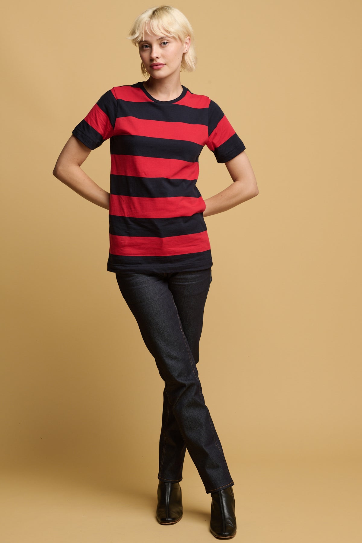 Blonde female tood with one leg infront of the other with her arms resting behind her waist wearing wide stripe short sleeve t shirt in navy and red paired with straight leg jeans in indigo