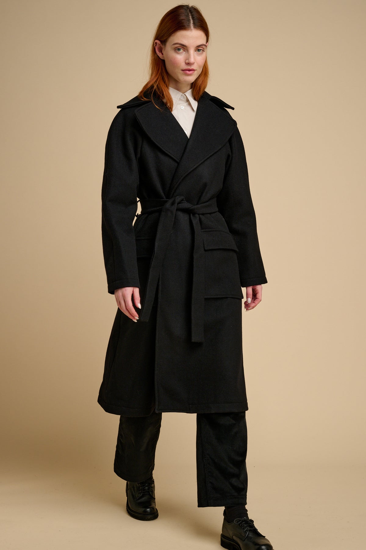 Woman wearing black wool coat with tie belt on beige background.