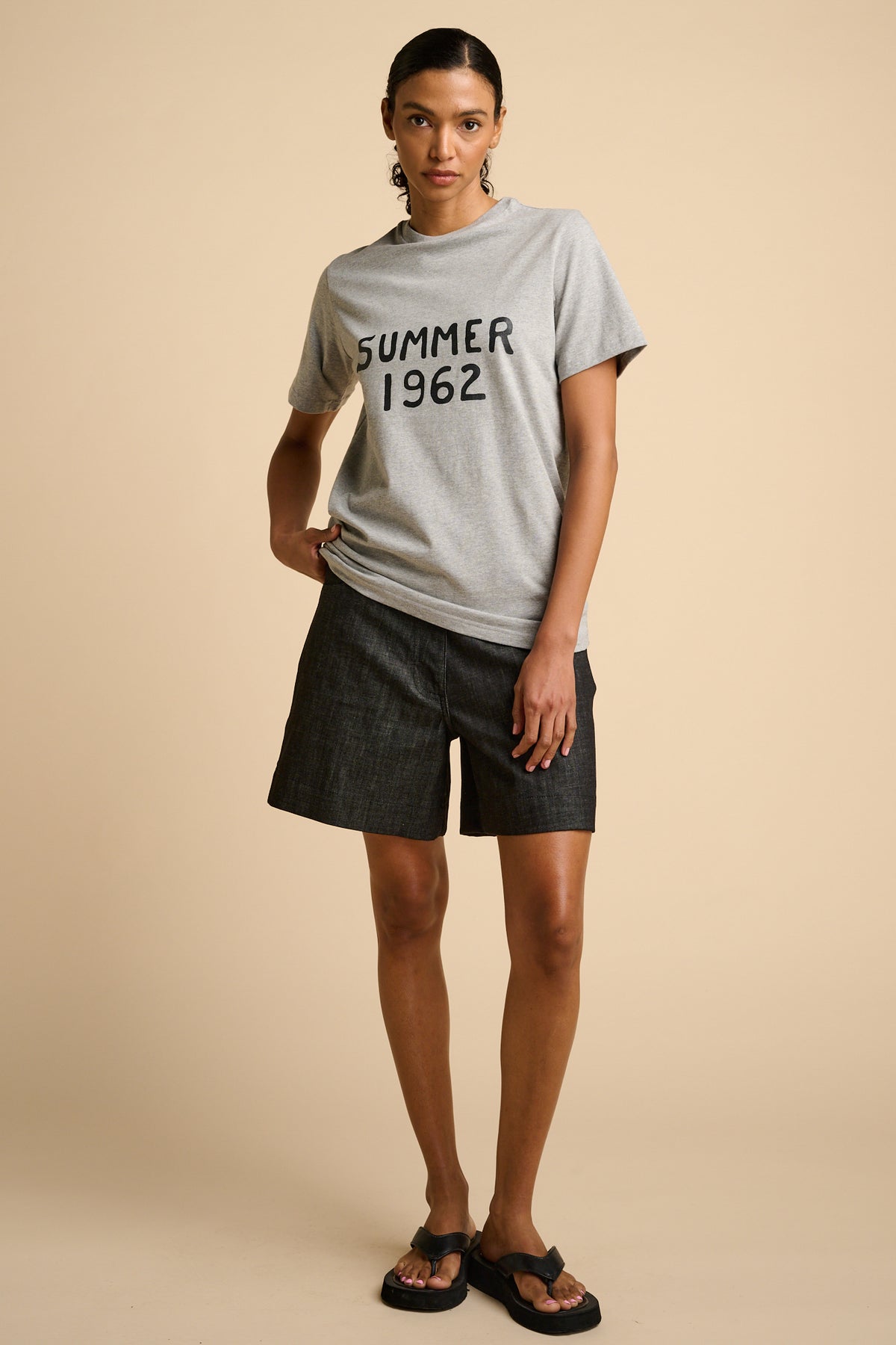 Woman wearing a grey t-shirt with 'Summer 1962' text with black shorts on a beige background.