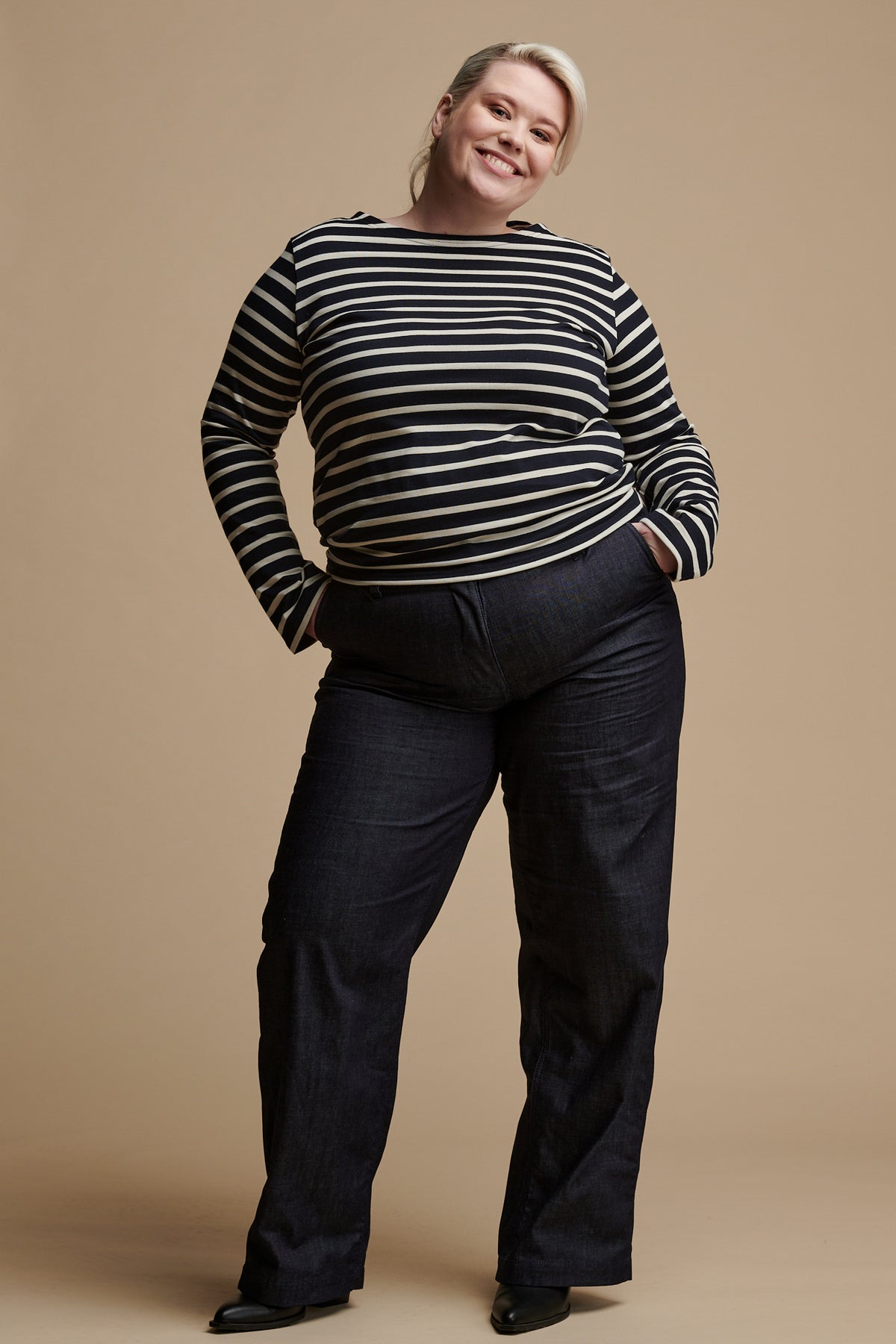 Smiling female wearing work trousers in indigo denim with both her hands in the front pockets paired with Breton top in navy-ecru.
