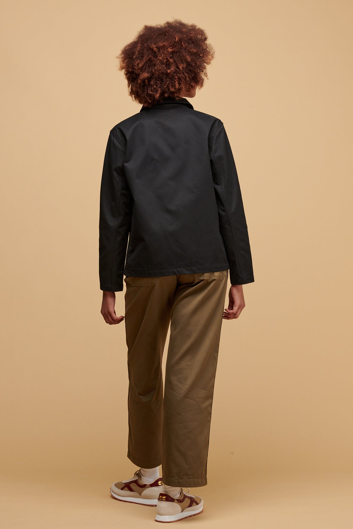 
            The back of female wearing chore jacket in black paired with work trousers in light olive and Walsh X Community Clothing Beacon trainers in neutral