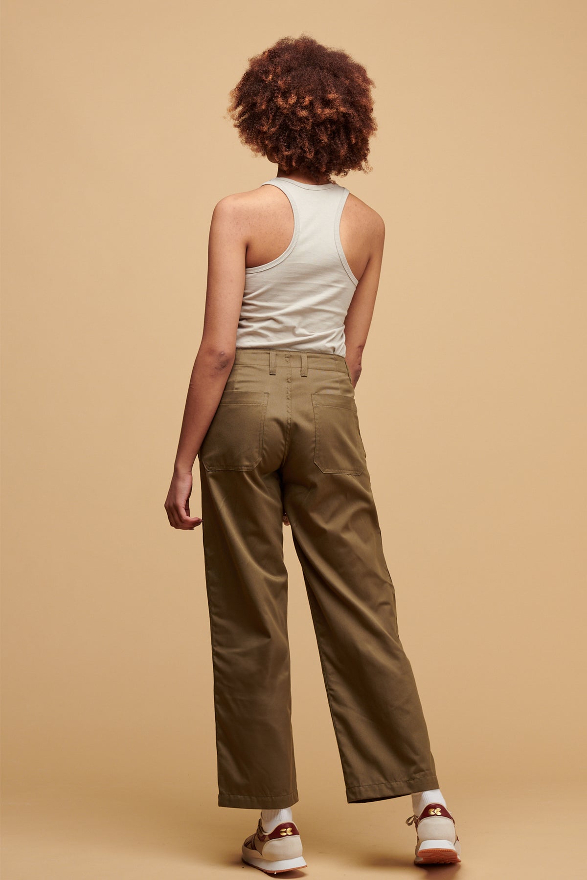 
            The back of female wearing work trousers in light olive paired with racer back vest in stone tucked into trousers with Walsh X Community Clothing Beacon trainers in neutral 