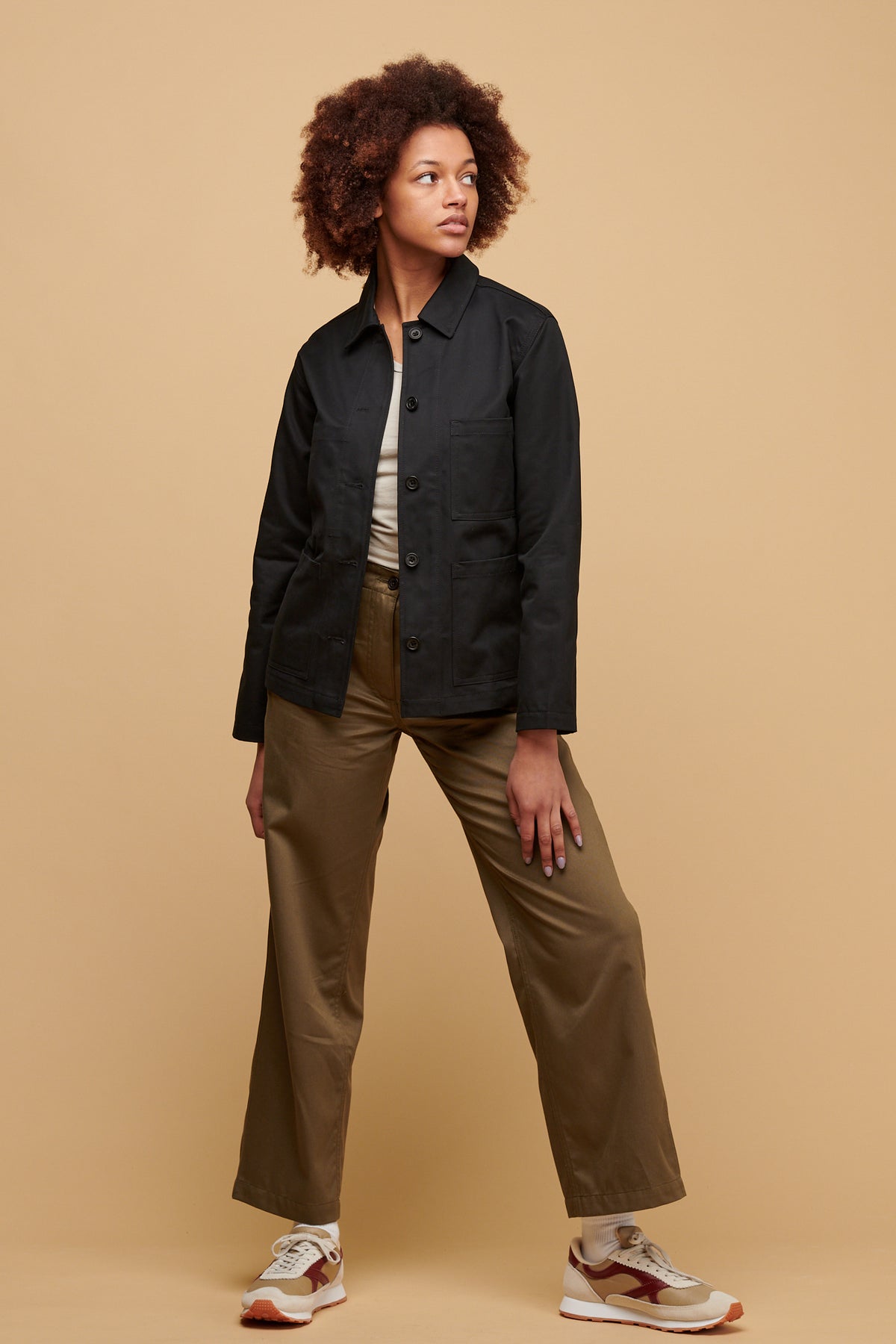 
            Female wearing chore jacket in black unbuttoned over racer back vest in stone paired with work trousers in light olive and Walsh X Community Clothing Beacon trainers in neutral.