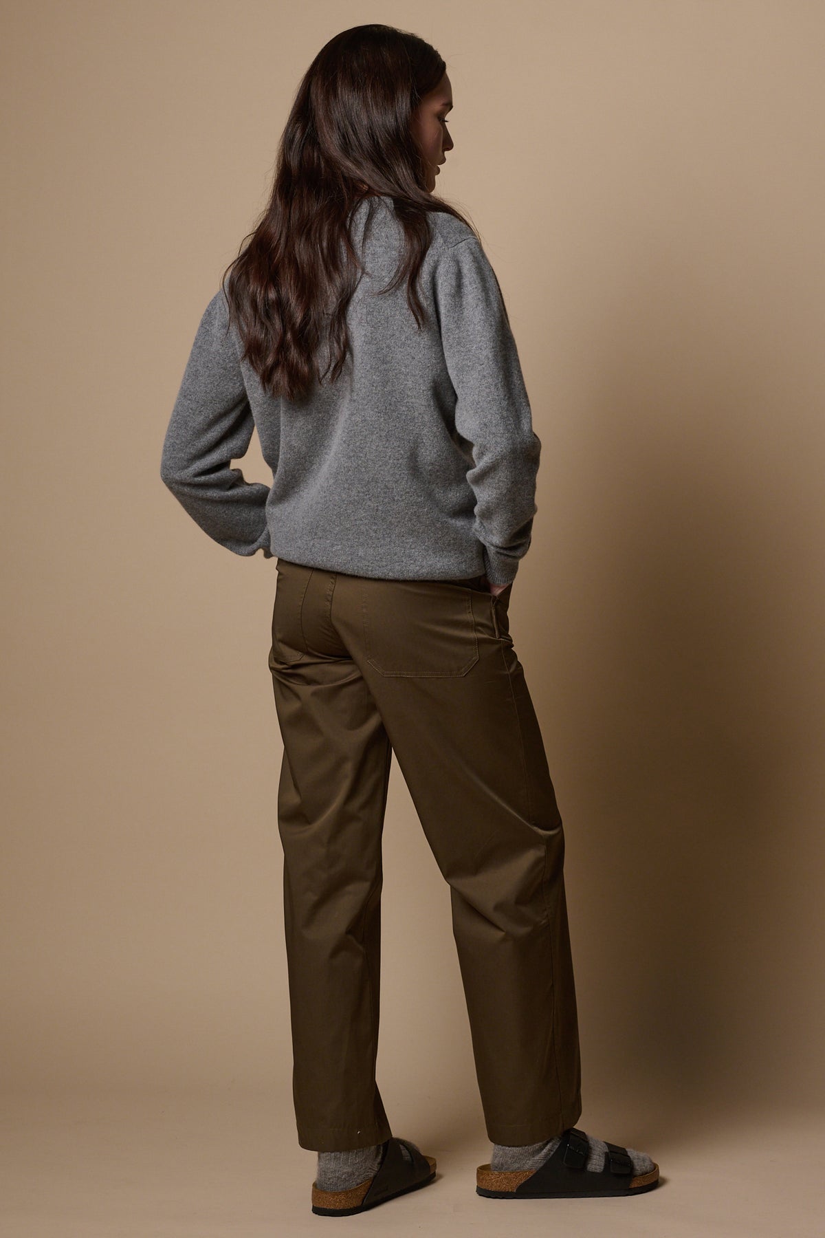 Full body image of the back of female wearing work trousers in olive with hands in front pockets.