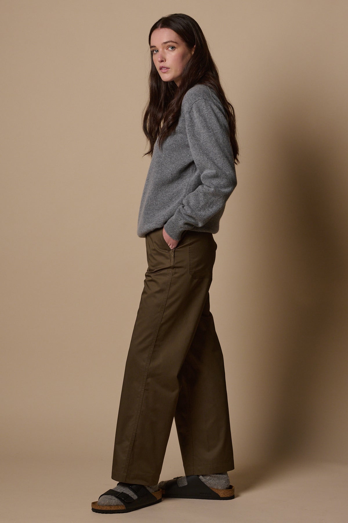 Full body shot of the side of brunette female wearing work trousers in olive with hand in front pocket. Worn with lambswool crew neck jumper