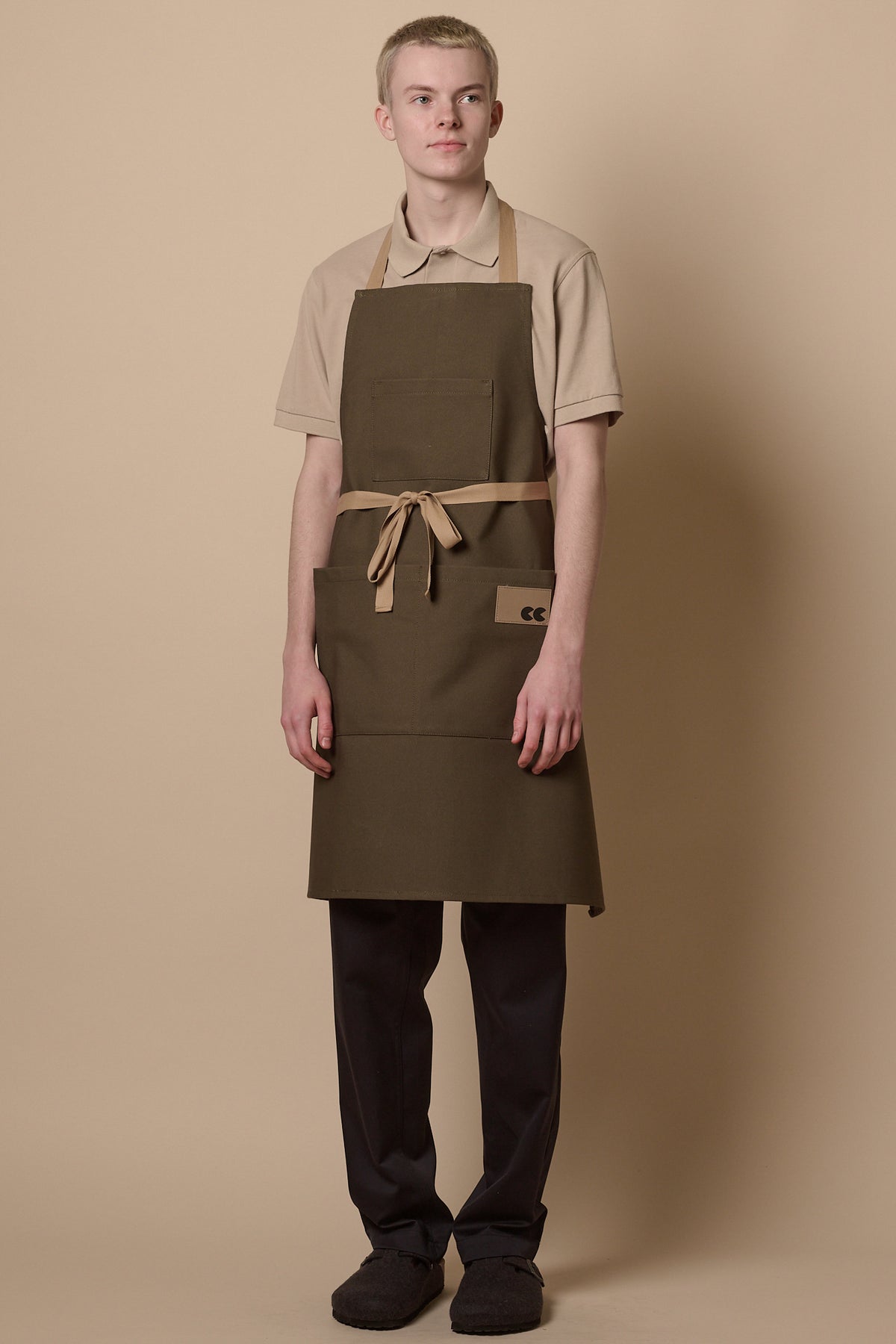
            Blonde male wearing apron in olive with beige ties tied at the front, three front pockets and CC logo patch