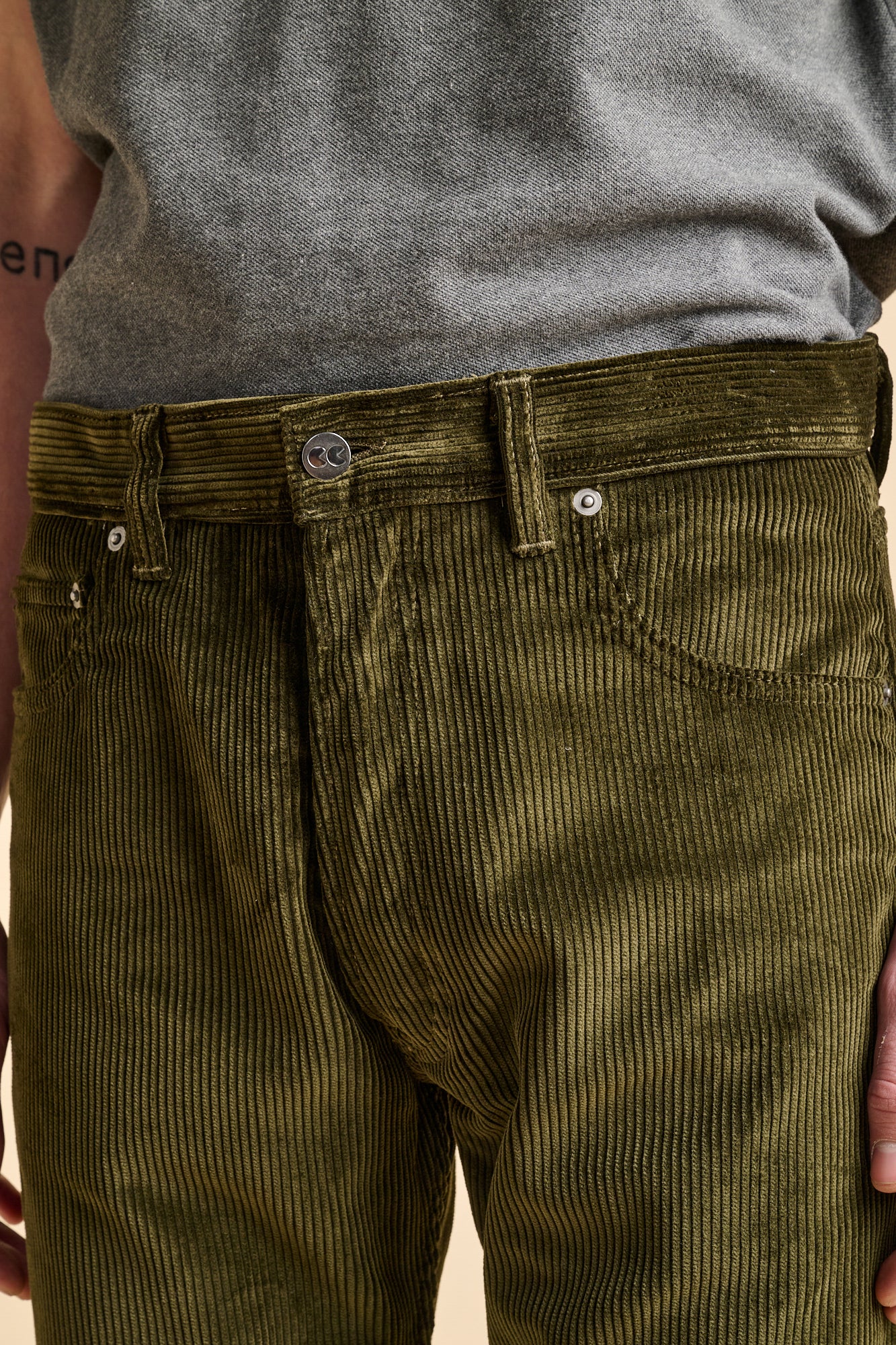 Close up the front of dark olive corduroy jeans with CC logo button fastening.