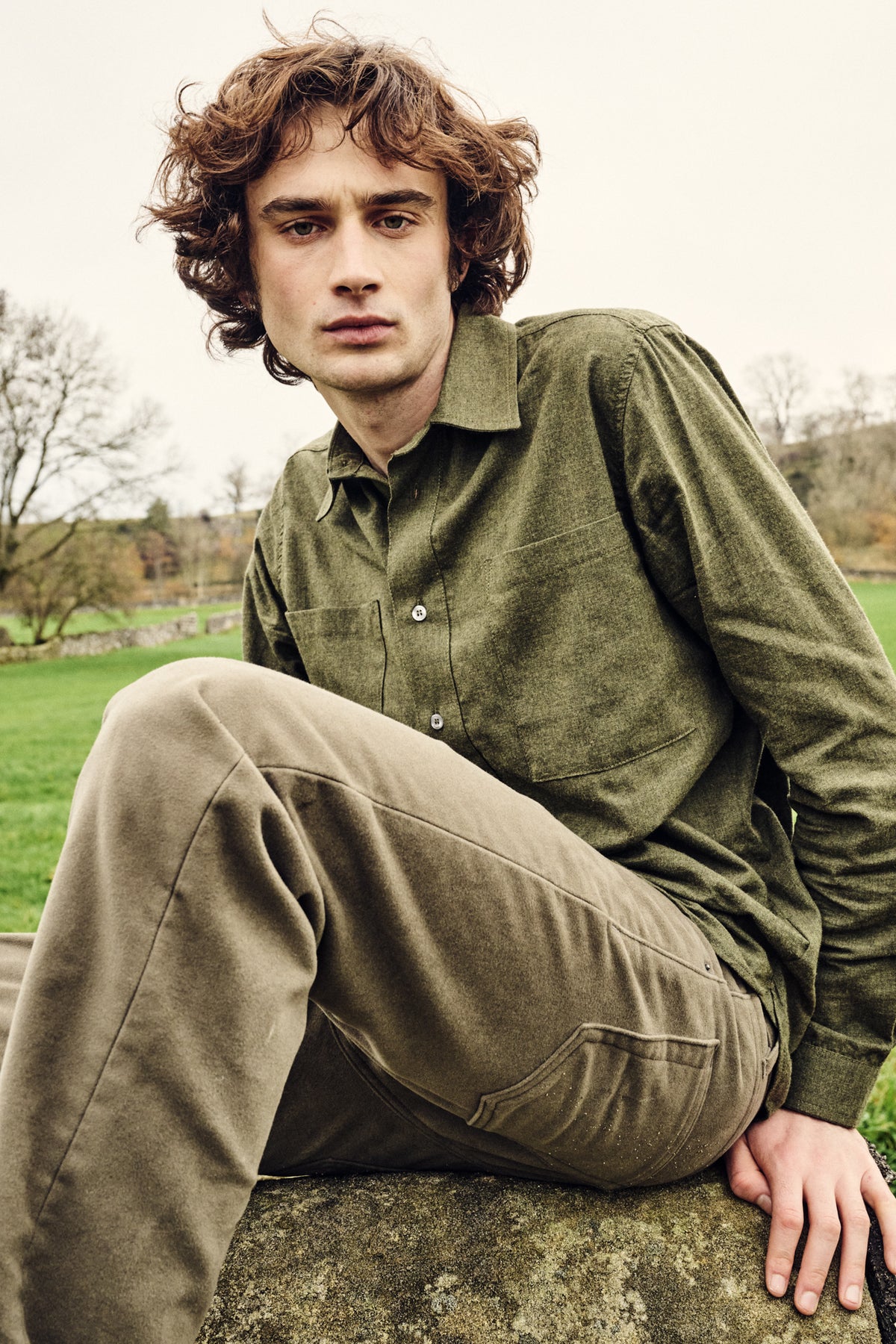 Man wearing green shirt and trousers sitting on a stone wall in a field with a natural landscape in the background