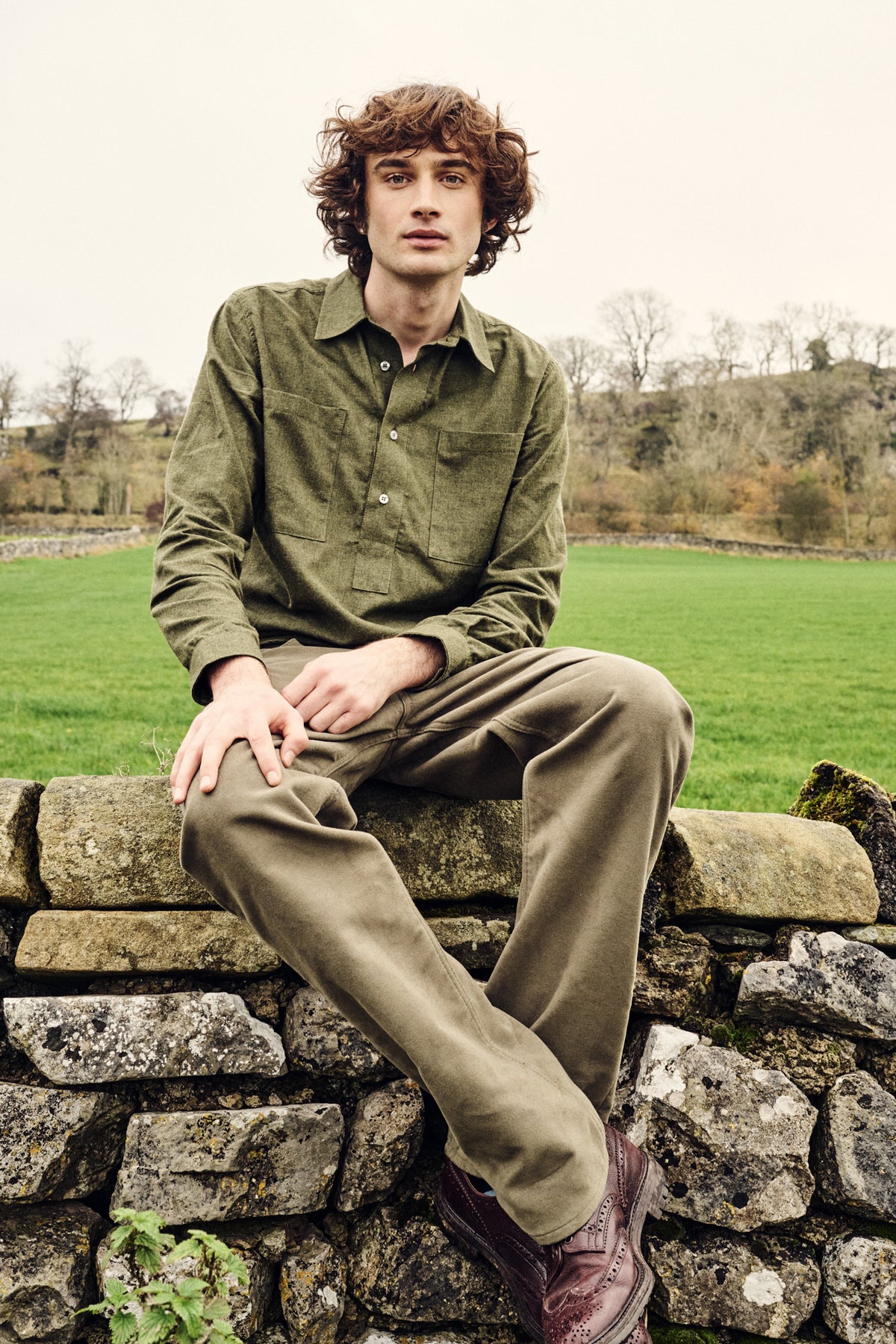 Man wearing green shirt and trousers sitting on a stone wall in a field with a natural landscape in the background