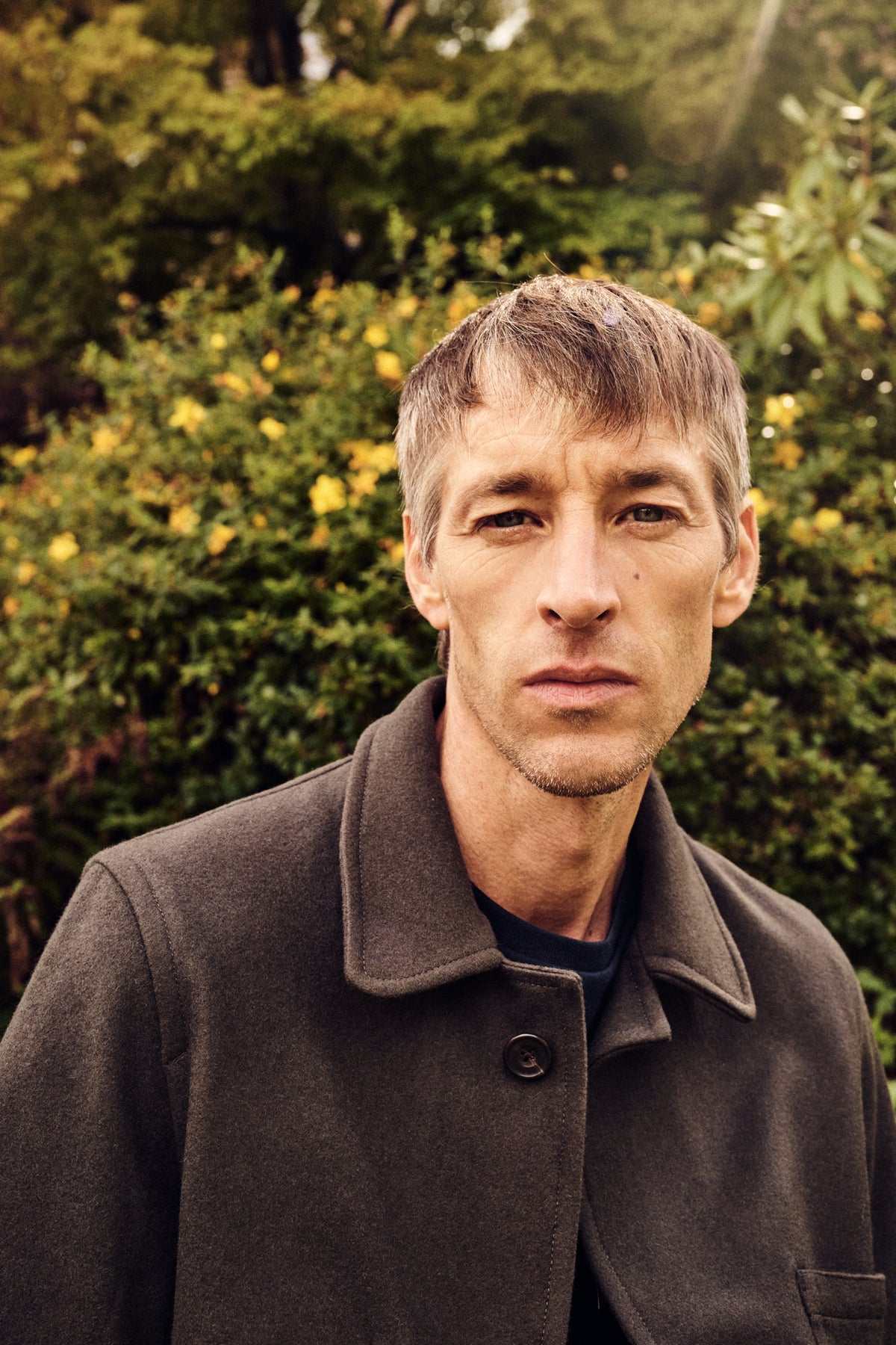 Man wearing a brown coat standing in front of green foliage