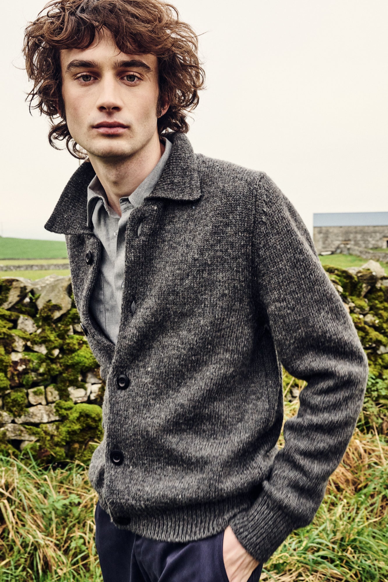 Man wearing a grey cardigan outside with stone walls and greenery.