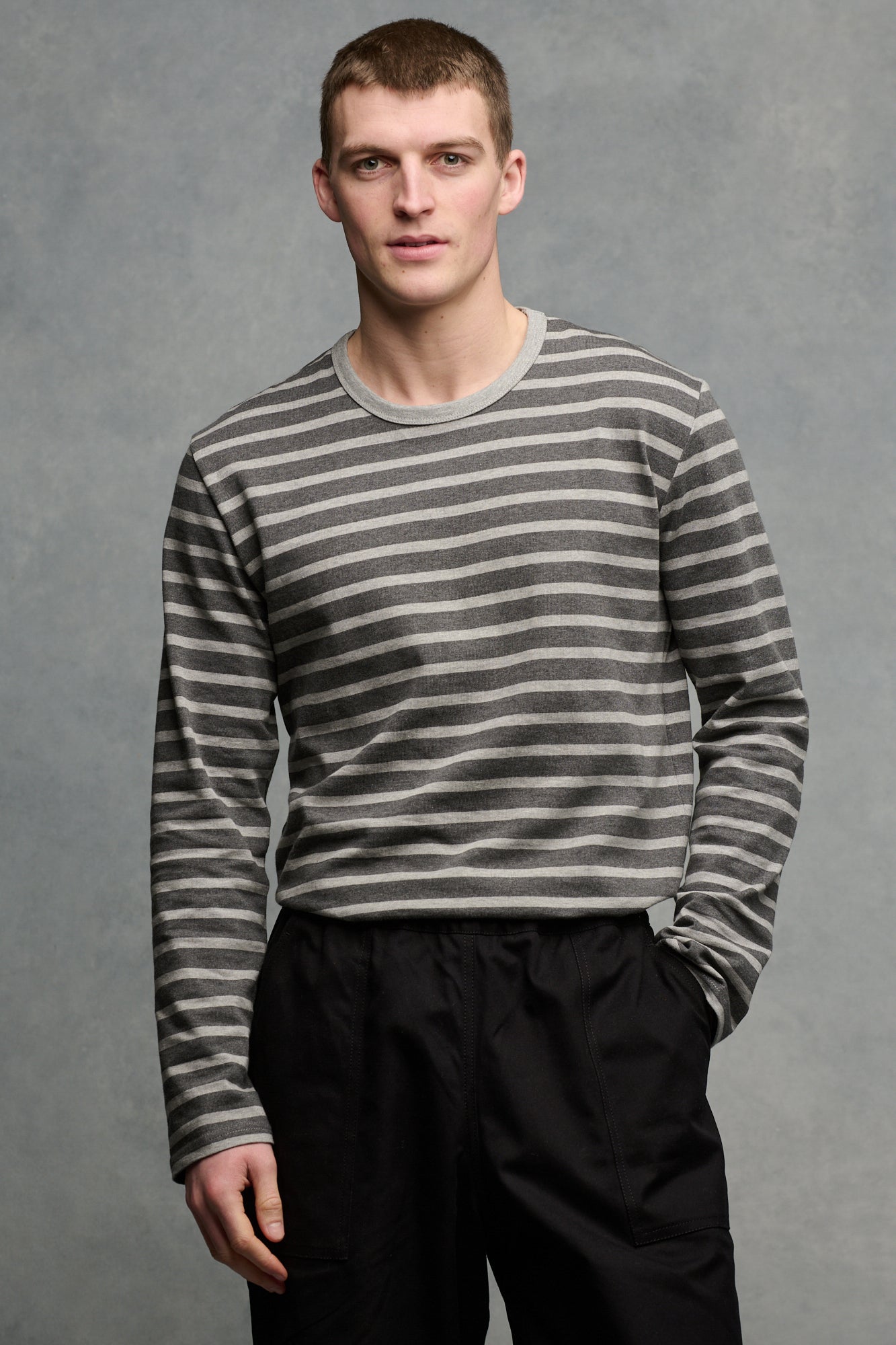 Thigh up image of the front of brunet male wearing charcoal marl/ grey marl tucked into cameraman pants in black 