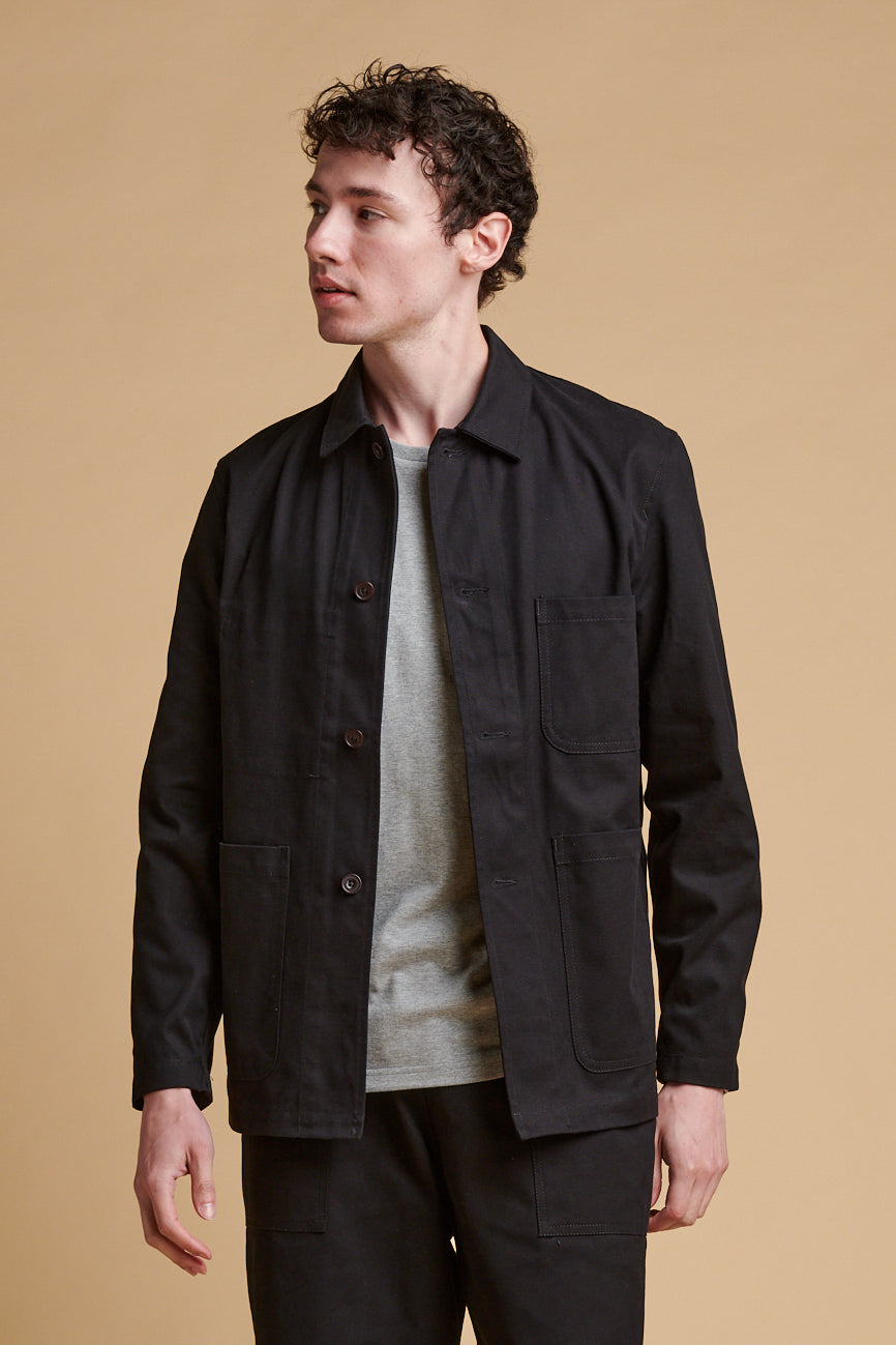 Male wearing chore jacket black unbuttoned over grey marl t shirt.