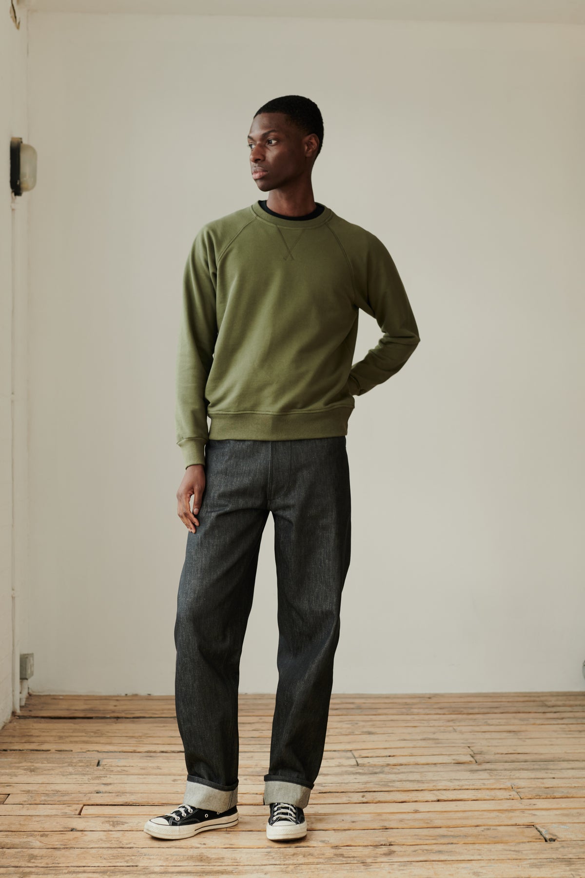 Black male wearing men's Chore jeans in black, wide leg and tapered fit with rolled hem, worn with olive green raglan sweatshirt