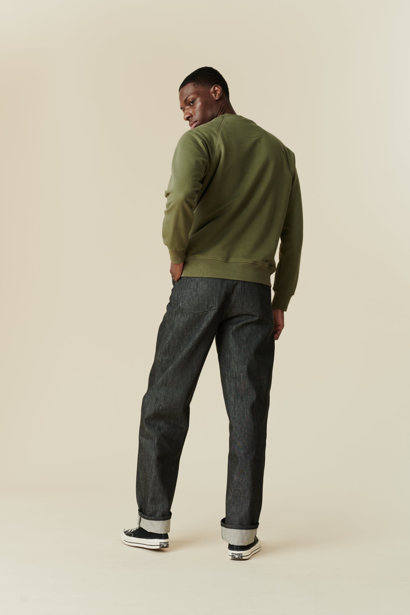 The back of black male wearing men's Chore jeans in black, wide leg and tapered fit with rolled hem, worn with olive green raglan sweatshirt