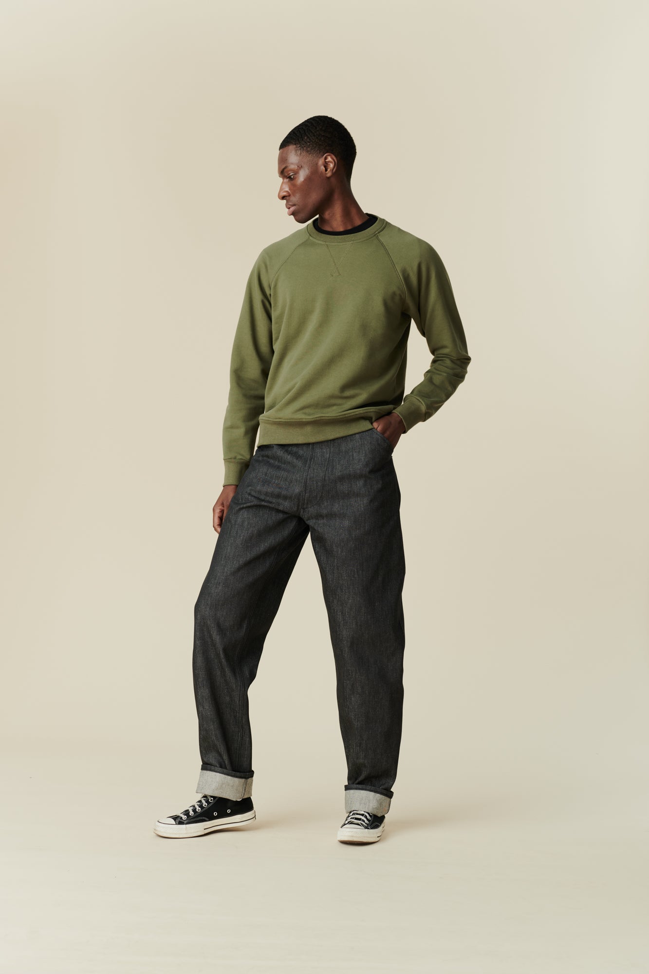 Full body image of black male wearing men's Chore jeans in black, wide leg and tapered fit with rolled hem, worn with olive green raglan sweatshirt