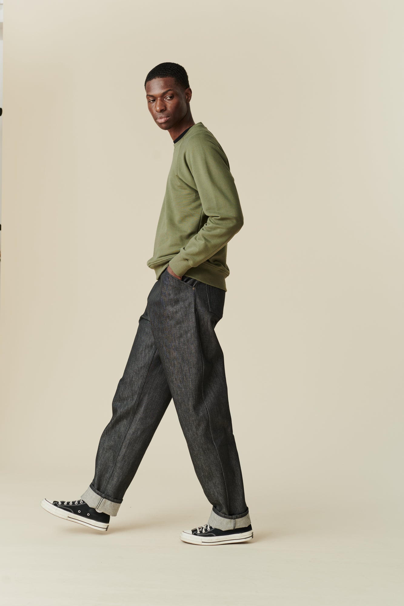 Side angle full body image of black male wearing men's Chore jeans in black, wide leg and tapered fit with rolled hem, worn with olive green raglan sweatshirt