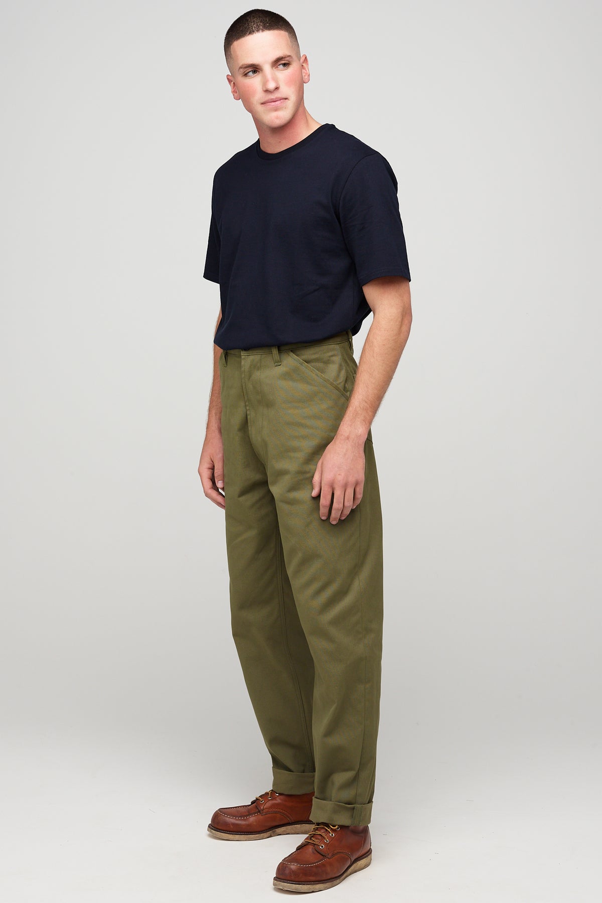 Brunette, white male wearing chore trousers in olive with short sleeve t shirt in navy tucked into trousers.