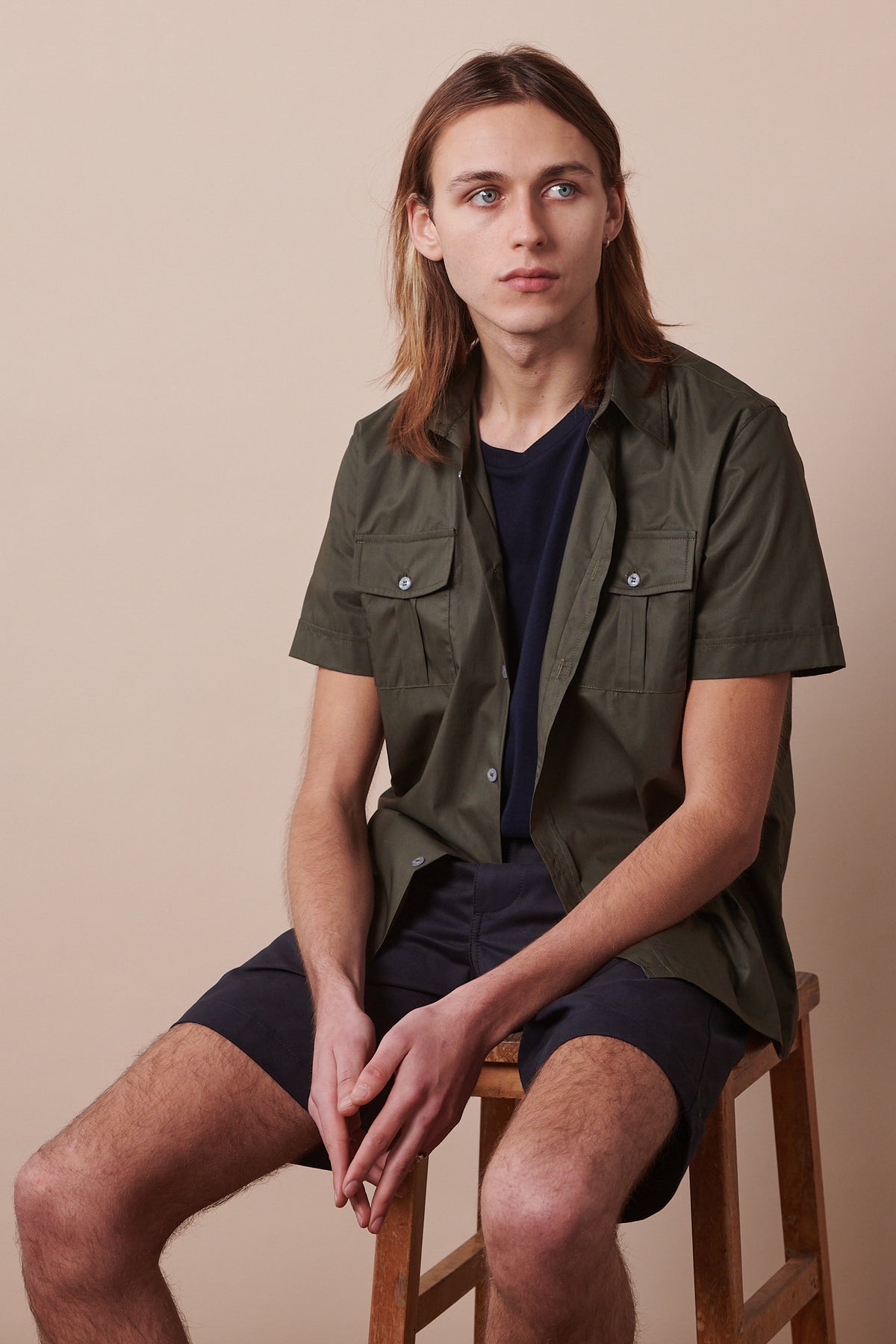 Male sat on stool wearing classic shorts in dark navy paired with tom military shirt in olive unbuttoned over navy t shirt