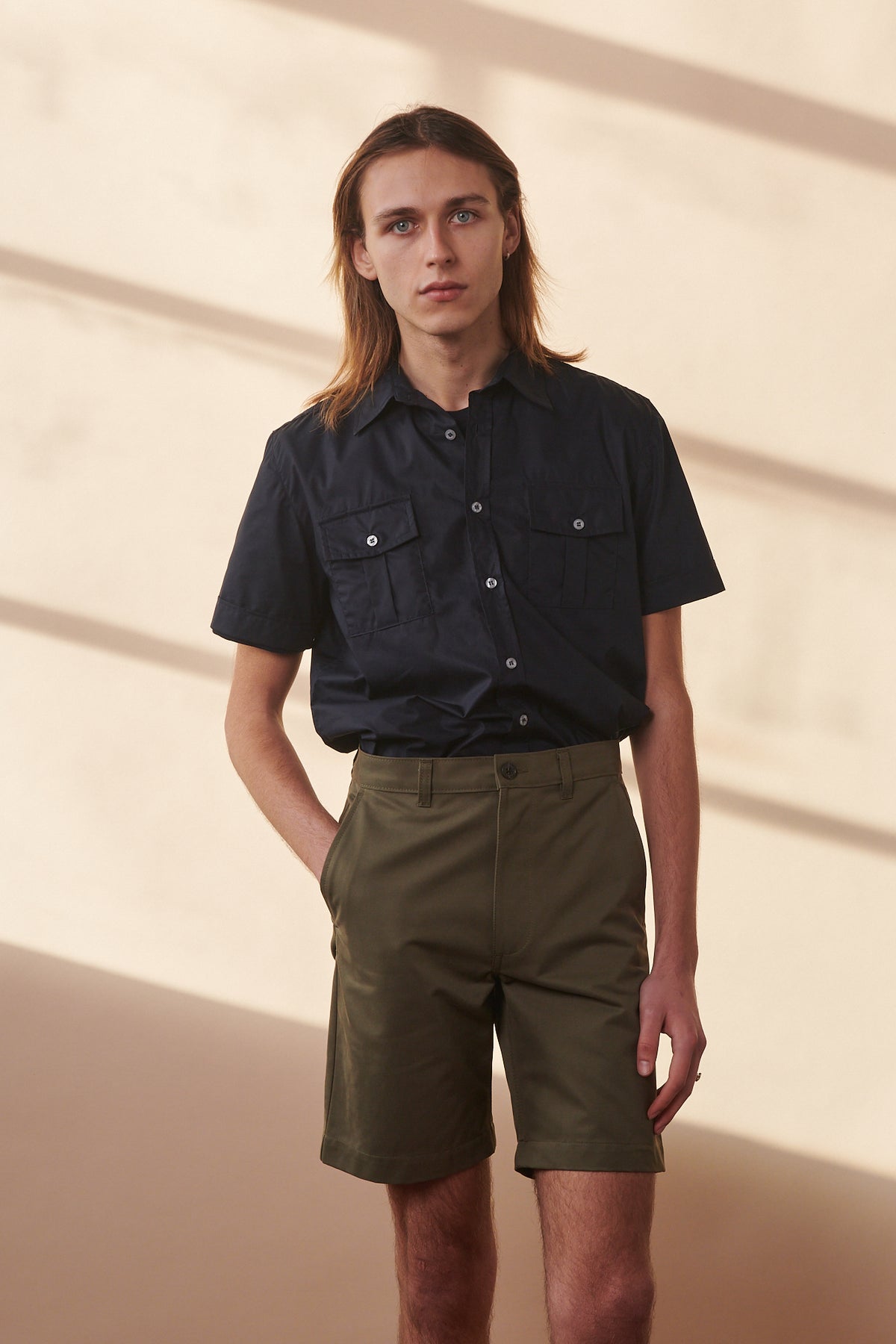 Male wearing classic shorts in olive with his hand in one of the front pockets paired with navy military shirt buttoned and tucked into shorts